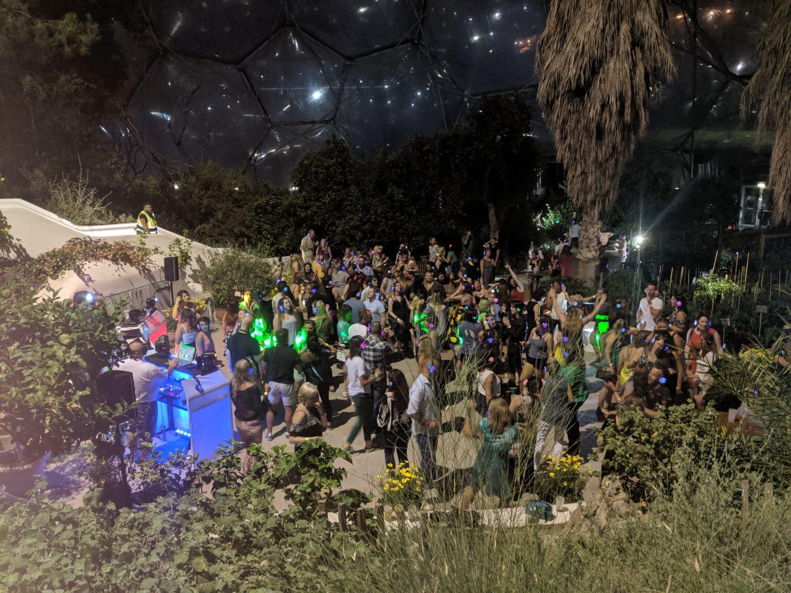 Nighttime party in a dome with a crowd dancing; colorful lights, DJ booth, and foliage are visible.