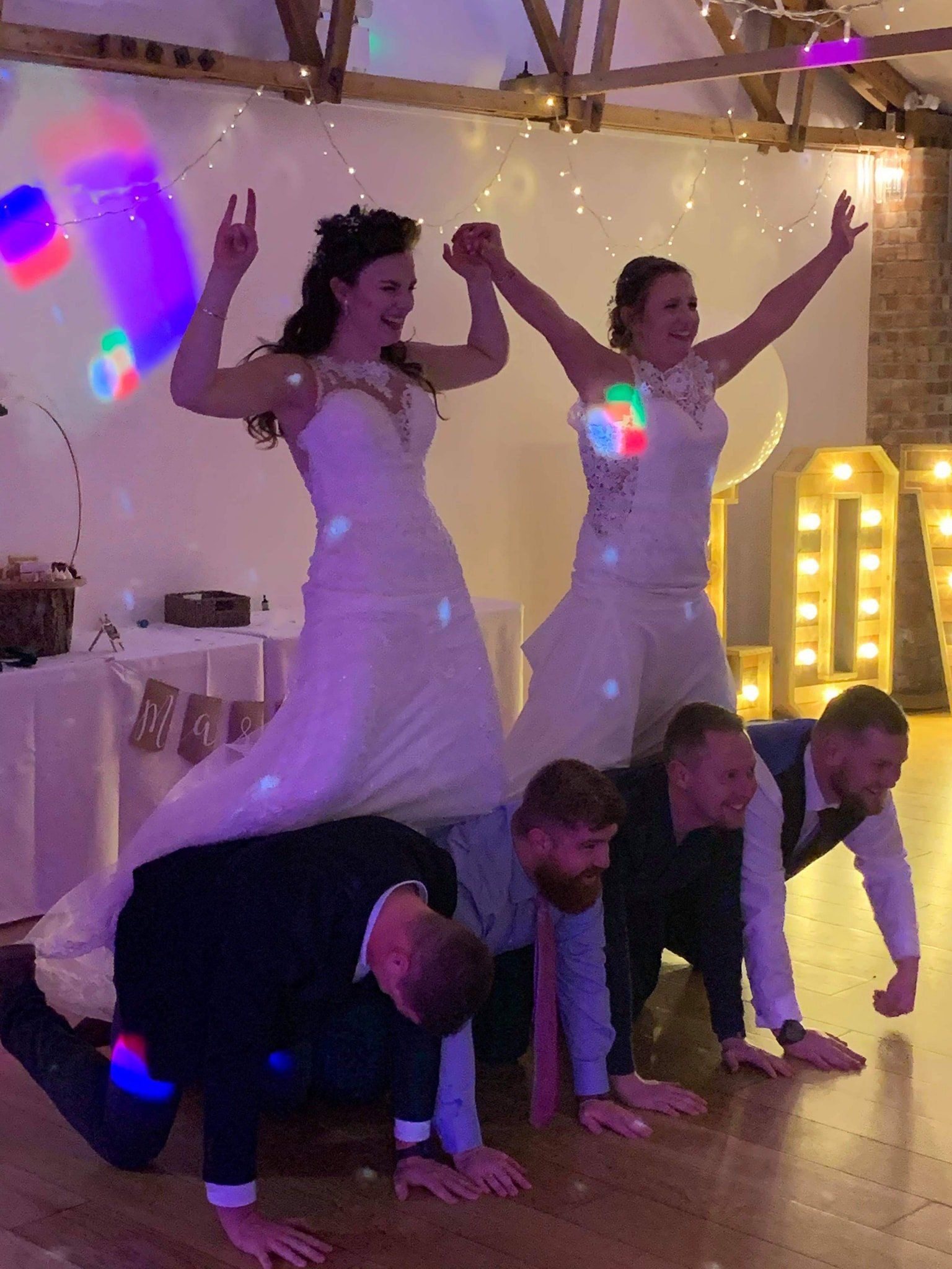 Two brides stand atop four kneeling men, arms raised in celebration at a wedding reception.