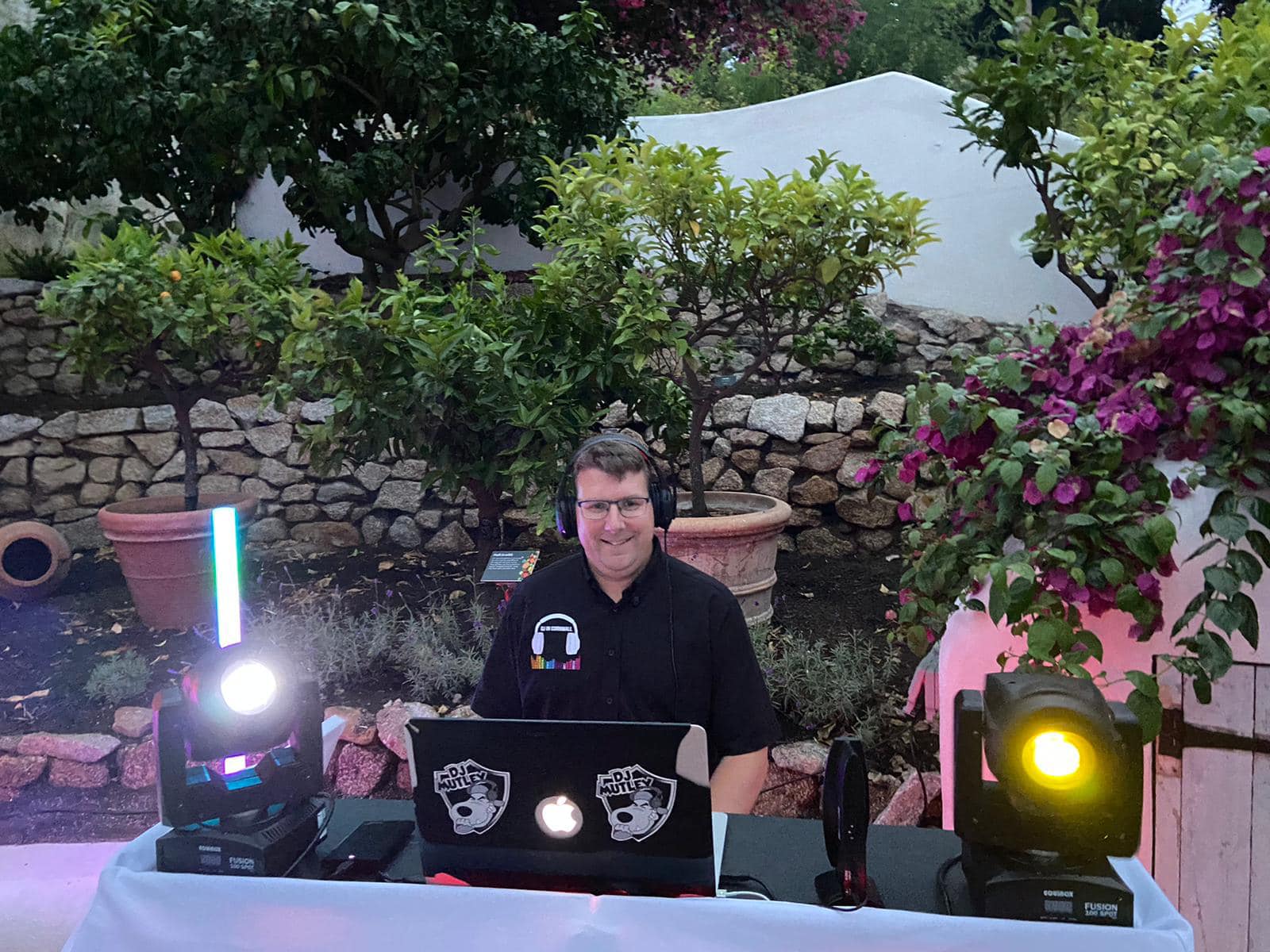 DJ at an outdoor event with lights, trees, and stone wall. Man in black shirt smiles at camera.