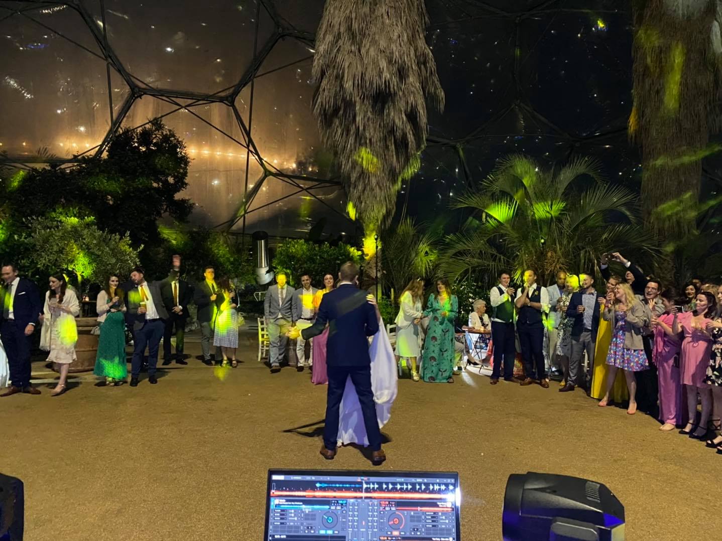 Wedding reception. People dance in a greenhouse-like setting. DJ in foreground. Nighttime.
