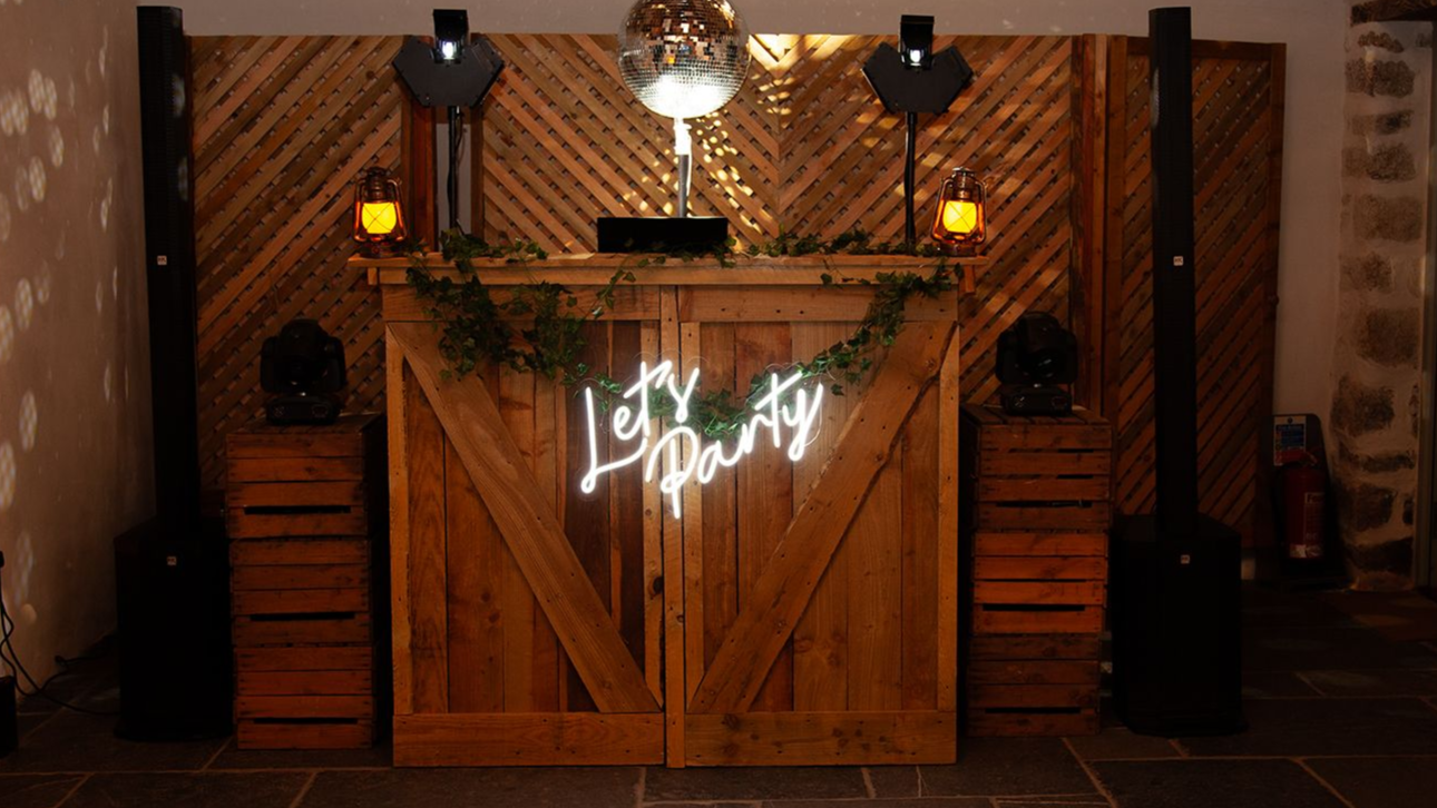 DJ setup with wooden crates, speakers, lights, and a wooden DJ booth, in a room with stone walls and a large window.