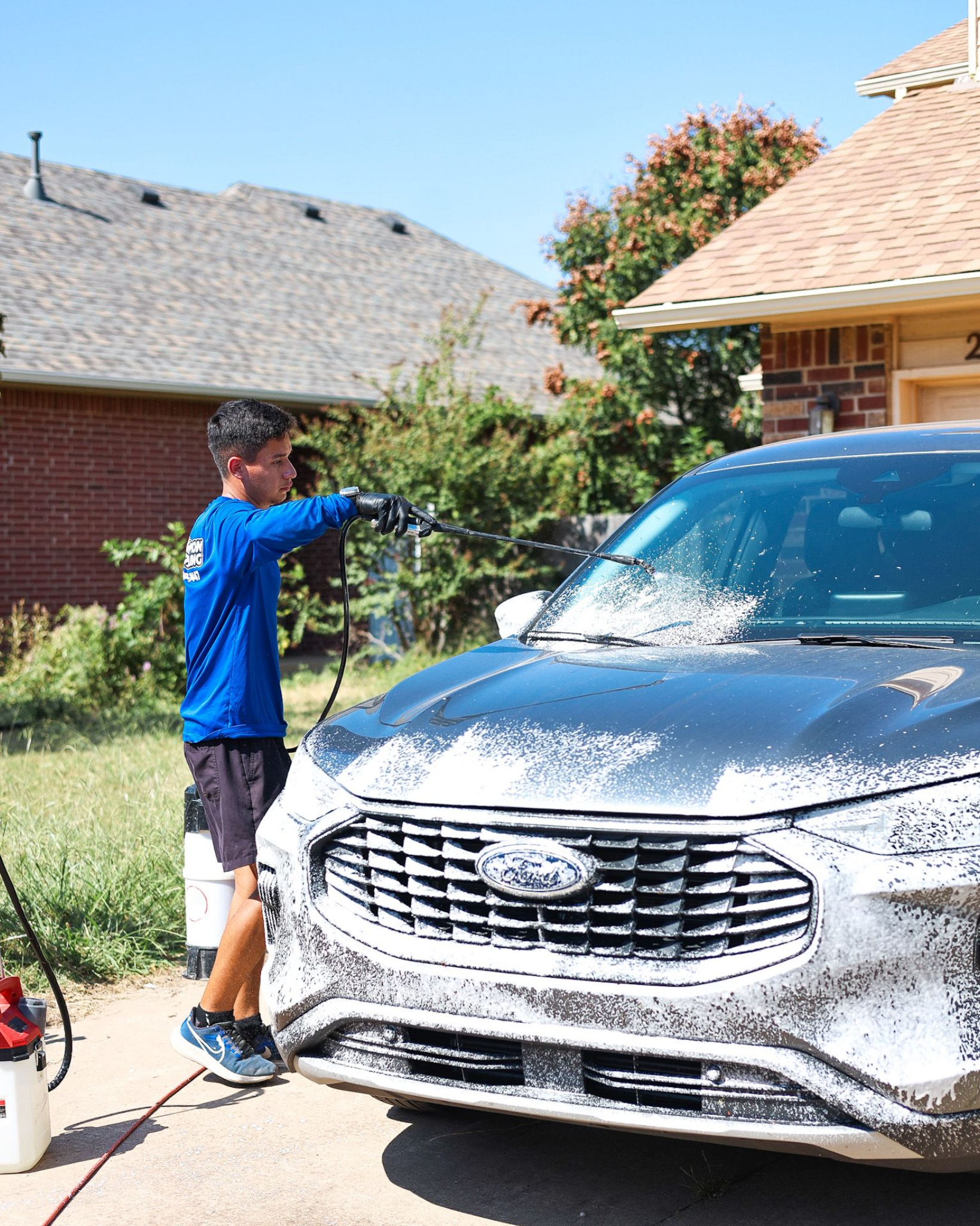 Person spray-washing a white SUV in a suburban driveway.
