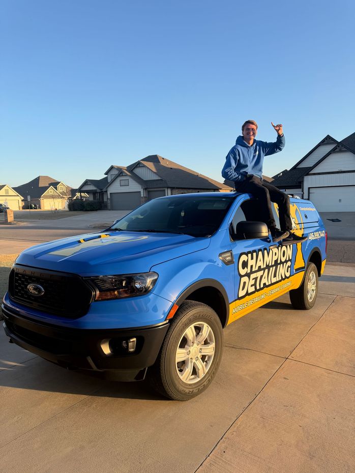 Blue-and-yellow detailing truck with a person waving from the truck bed in a suburban driveway at sunset