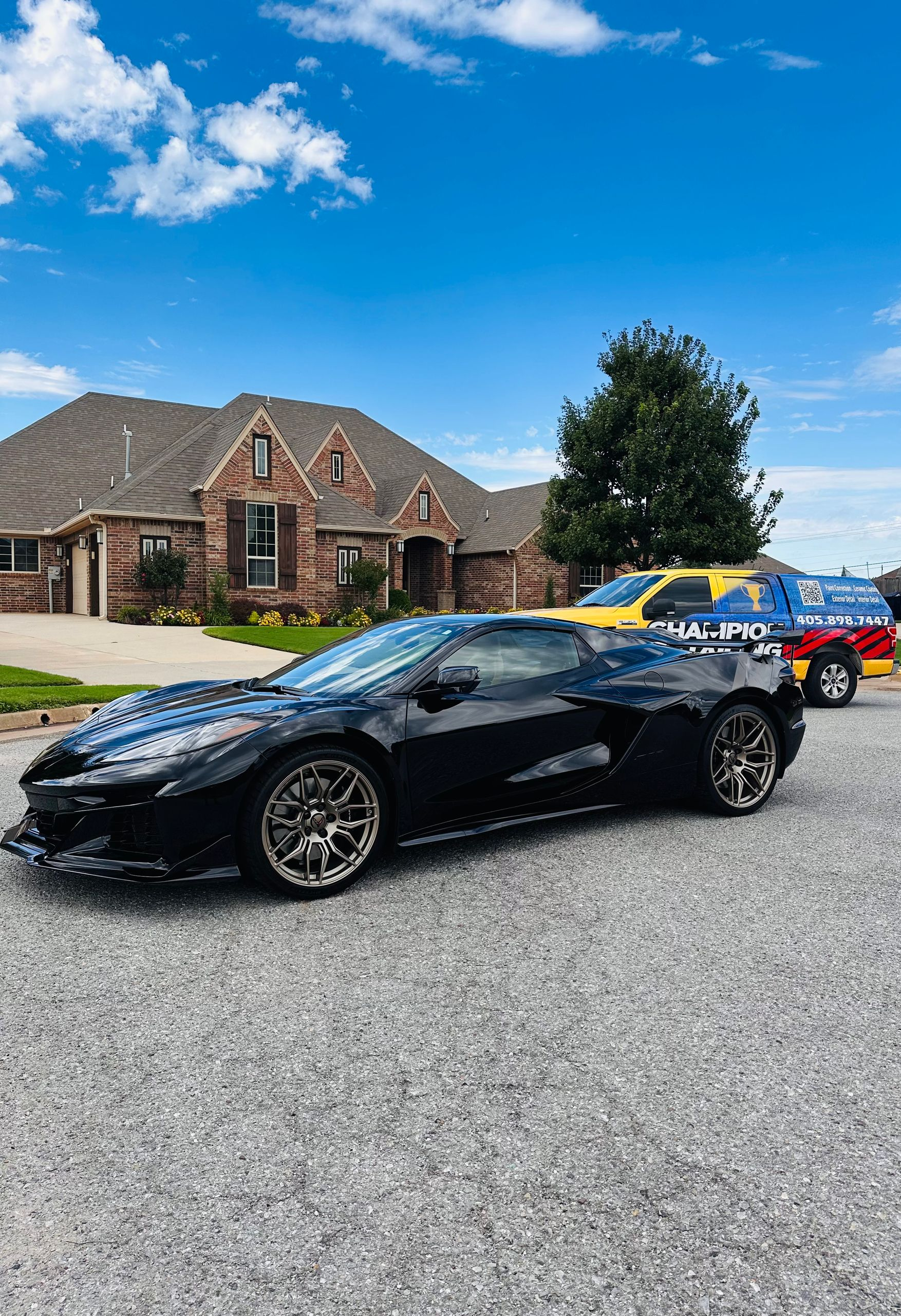 Black sports car parked on a gravel driveway in front of a brick house under a blue sky