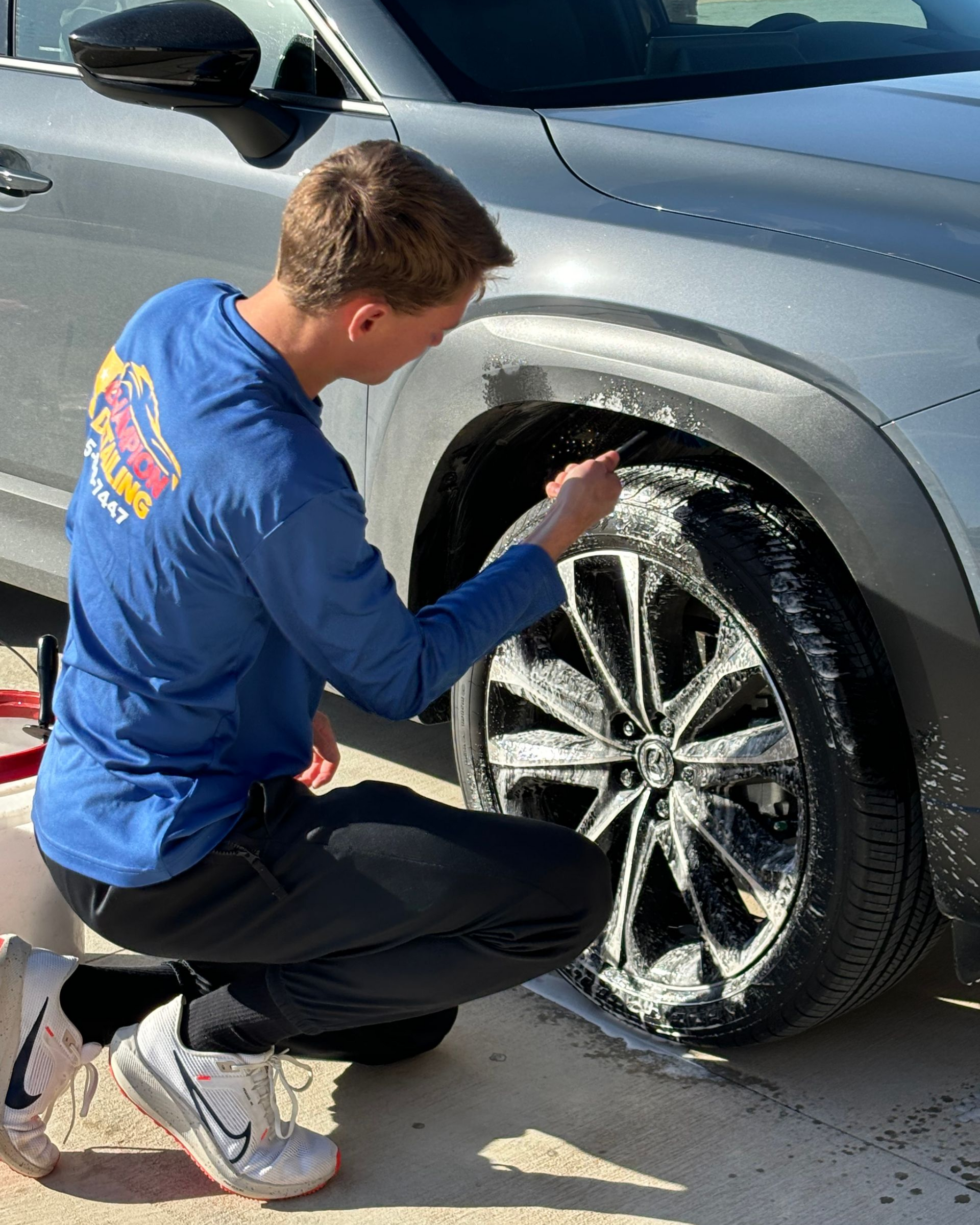 Person kneels beside a silver SUV, washing the front wheel with soap and a brush.