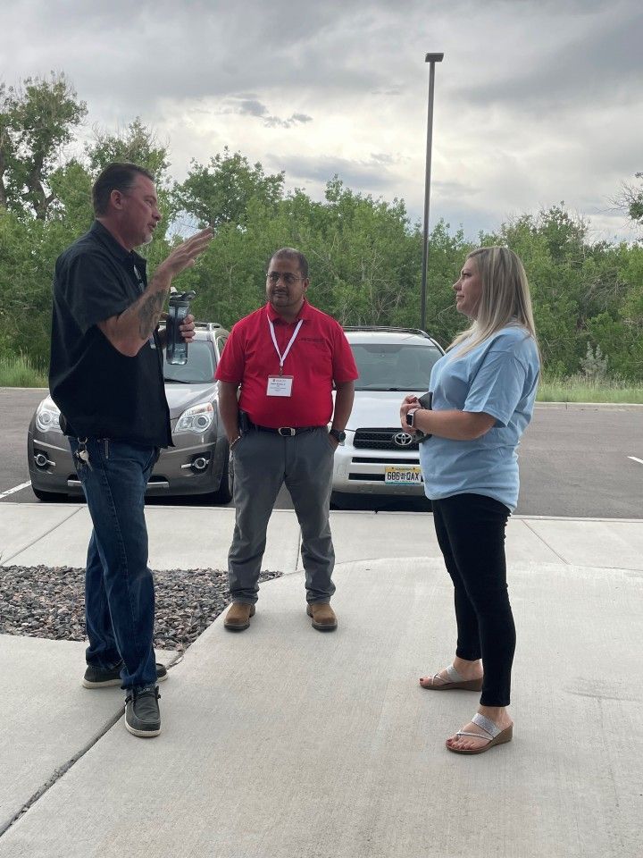 Three people in conversation outside; man gesturing, other man with red lanyard, woman listening.