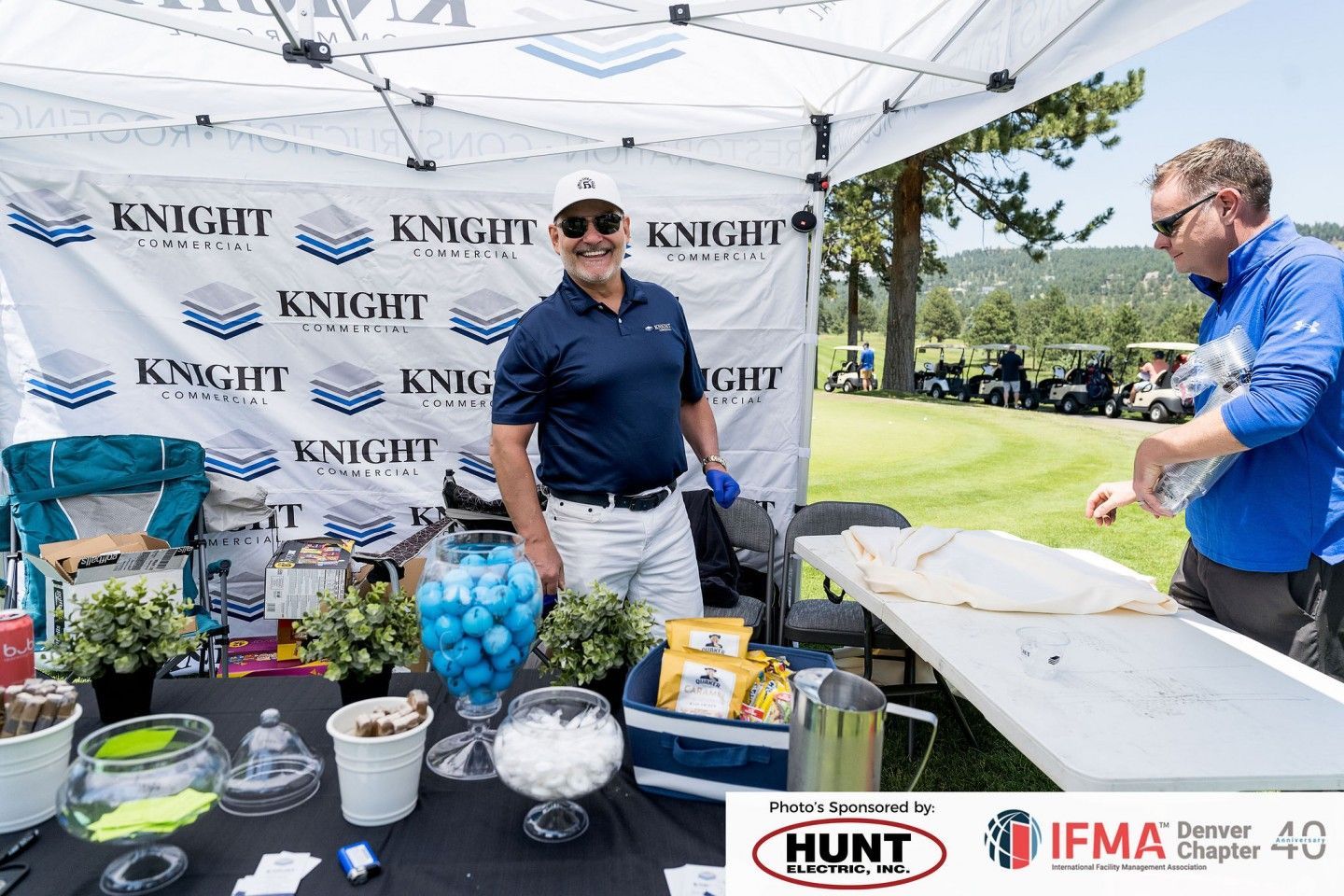 Man smiles at a table under a white tent at a golf event. Other people, golf carts, and a green course are in the background.