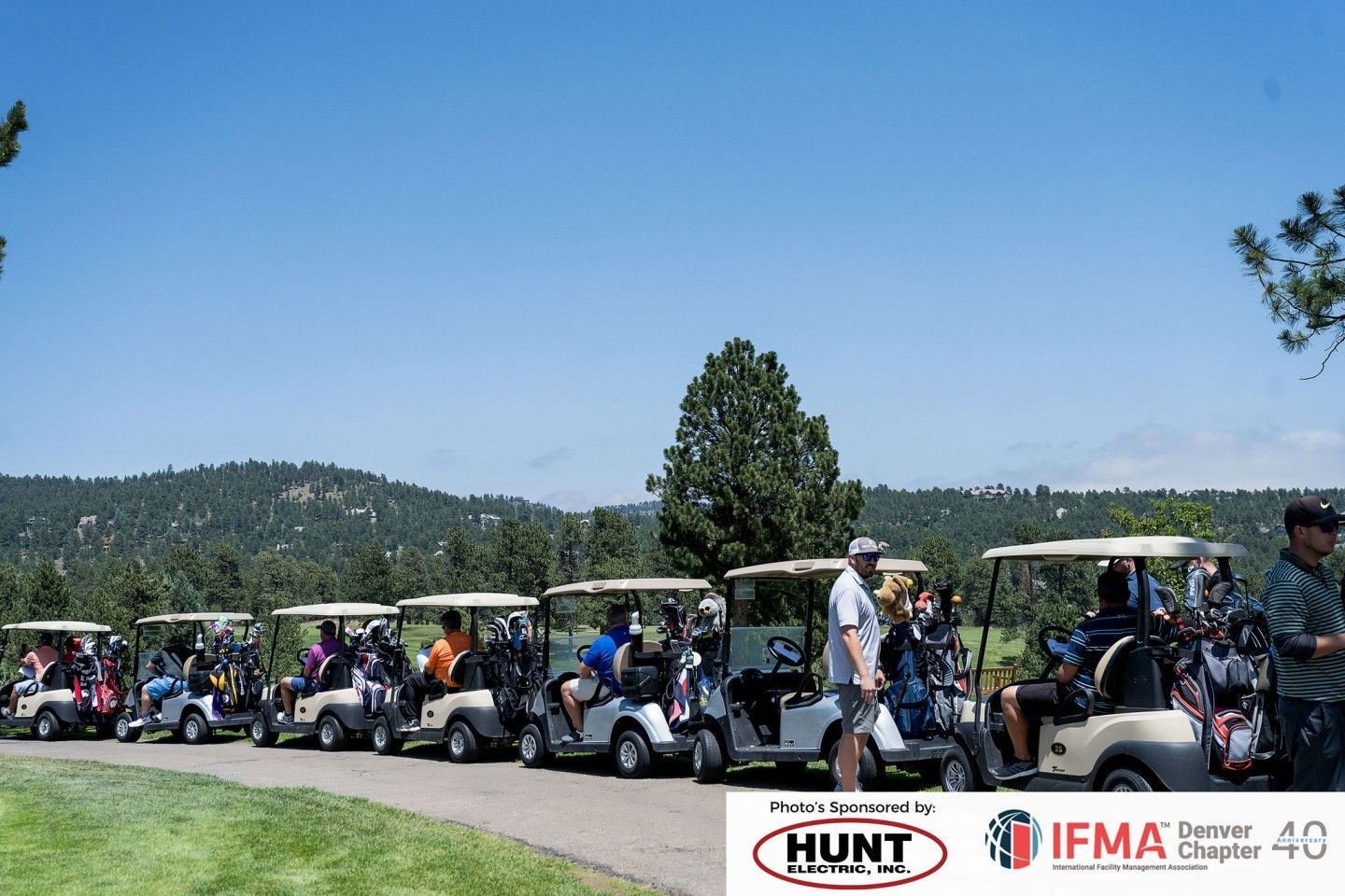 Golfers in golf carts lined up on a course, mountains in the background, under a blue sky.