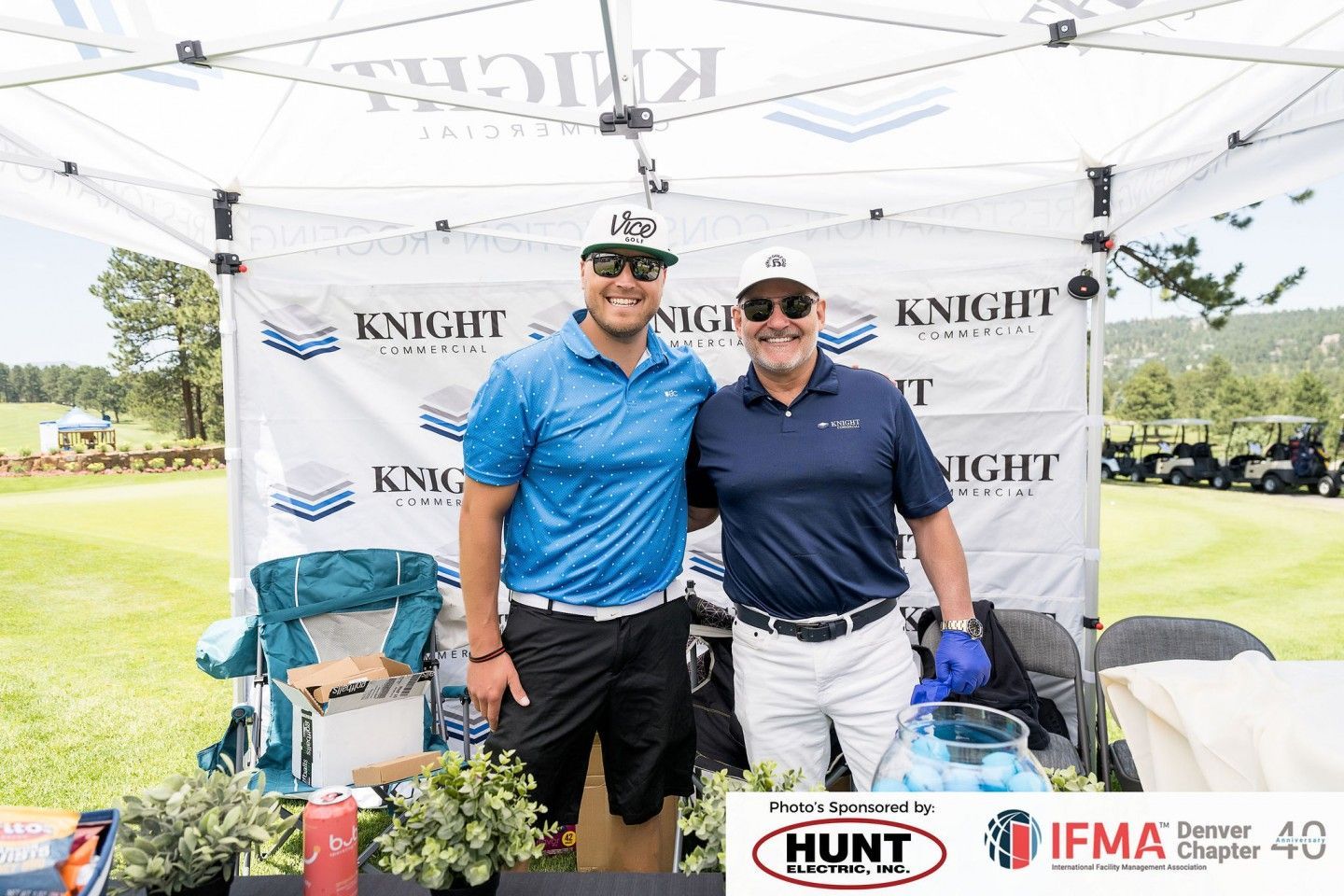Two men smiling under a tent with sponsor logos at an outdoor event.