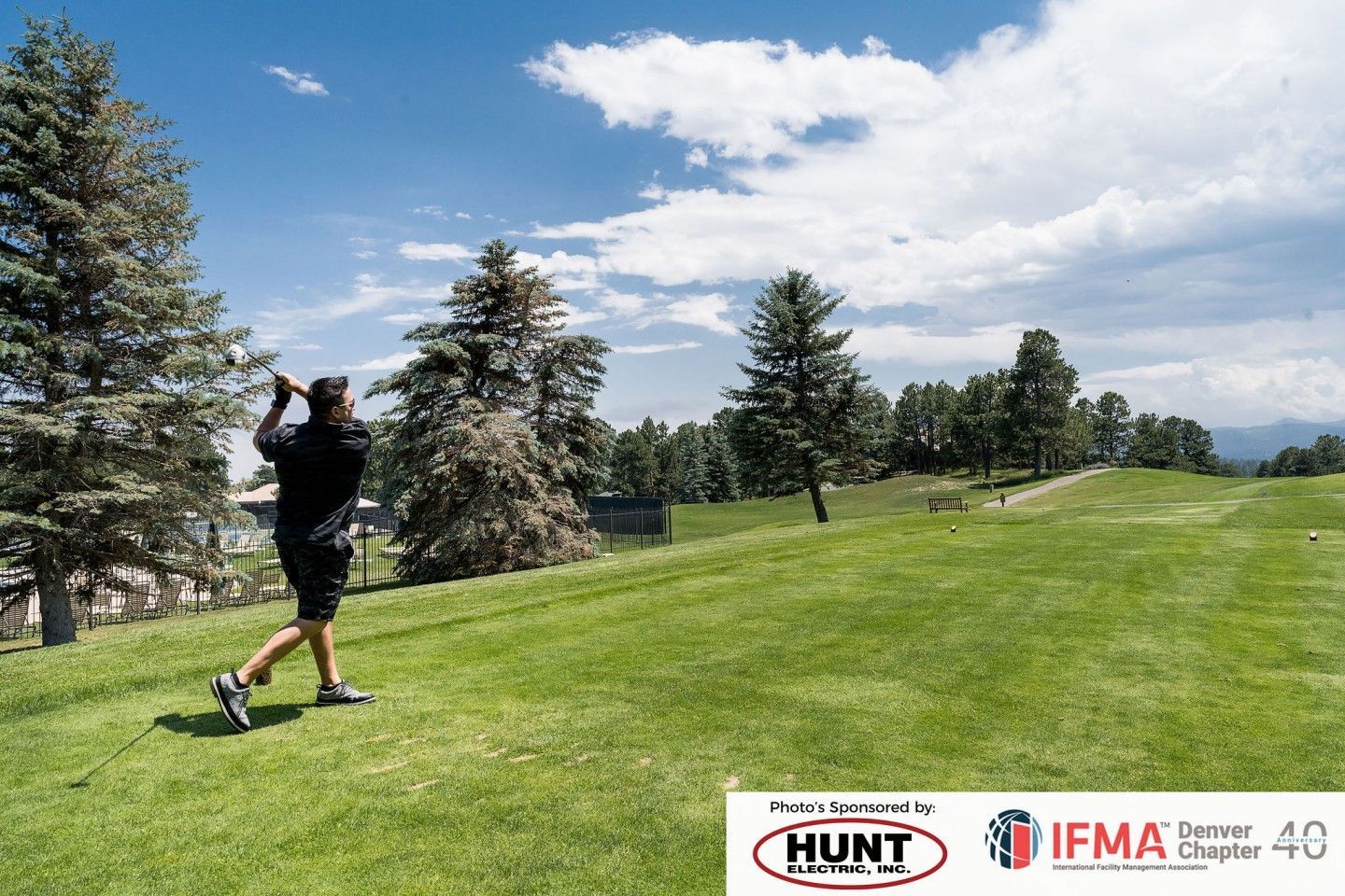 Man swings a golf club on a green course under a blue sky with fluffy clouds and trees in the background.