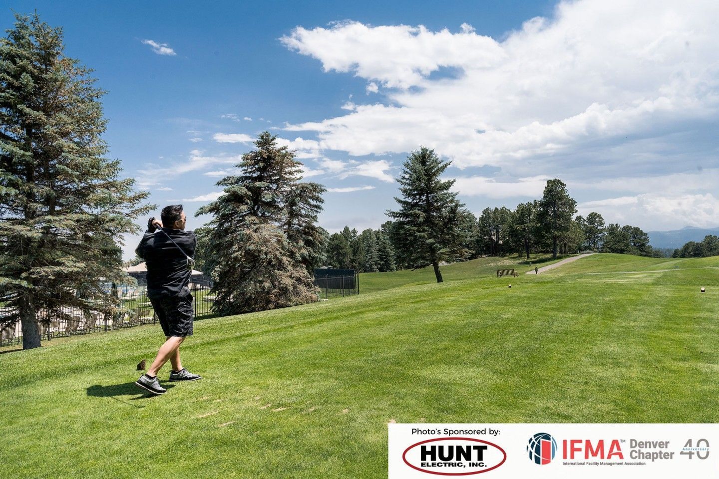 Man swings golf club on a green course under a blue sky with trees.