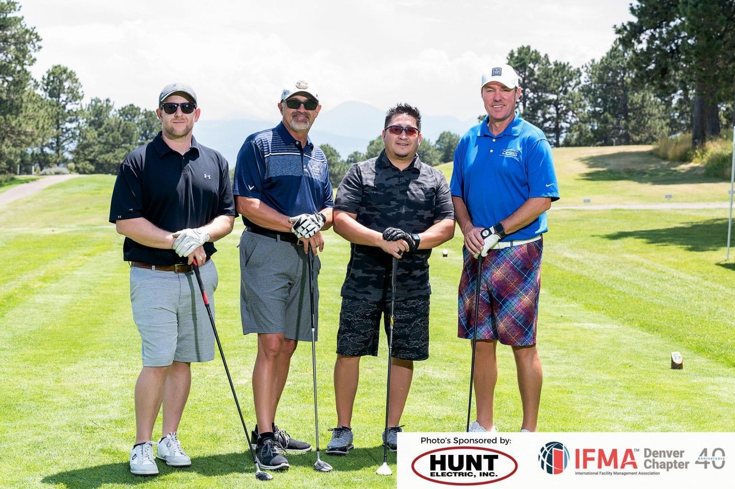 Four men in golf attire stand on a green, holding clubs. Mountains in background.