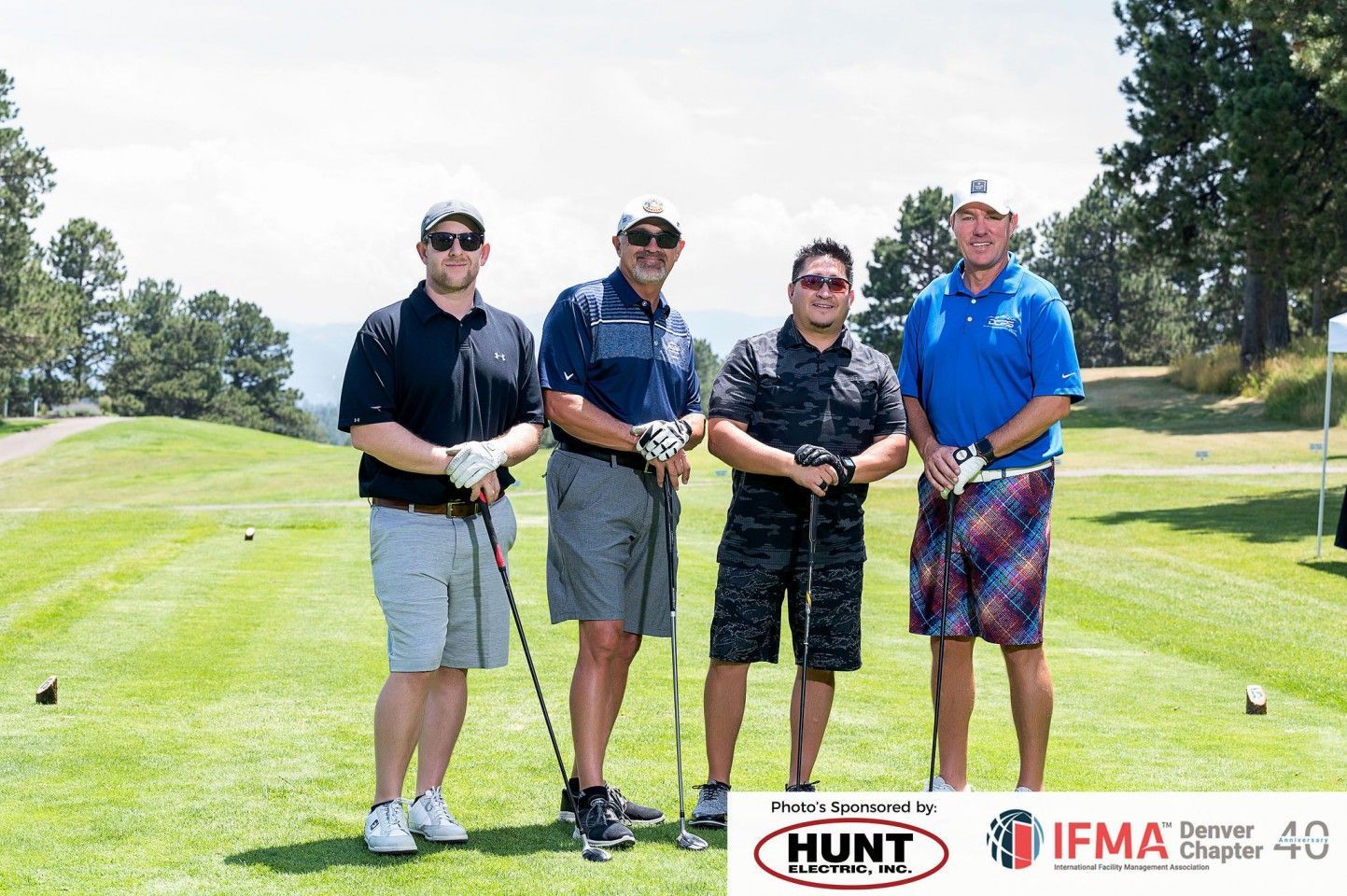 Four men on a golf course holding clubs, posing for a photo. Sunny day.