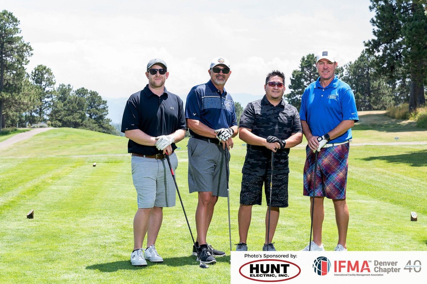 Four men with golf clubs on a golf course, posing for a photo under a sunny sky.