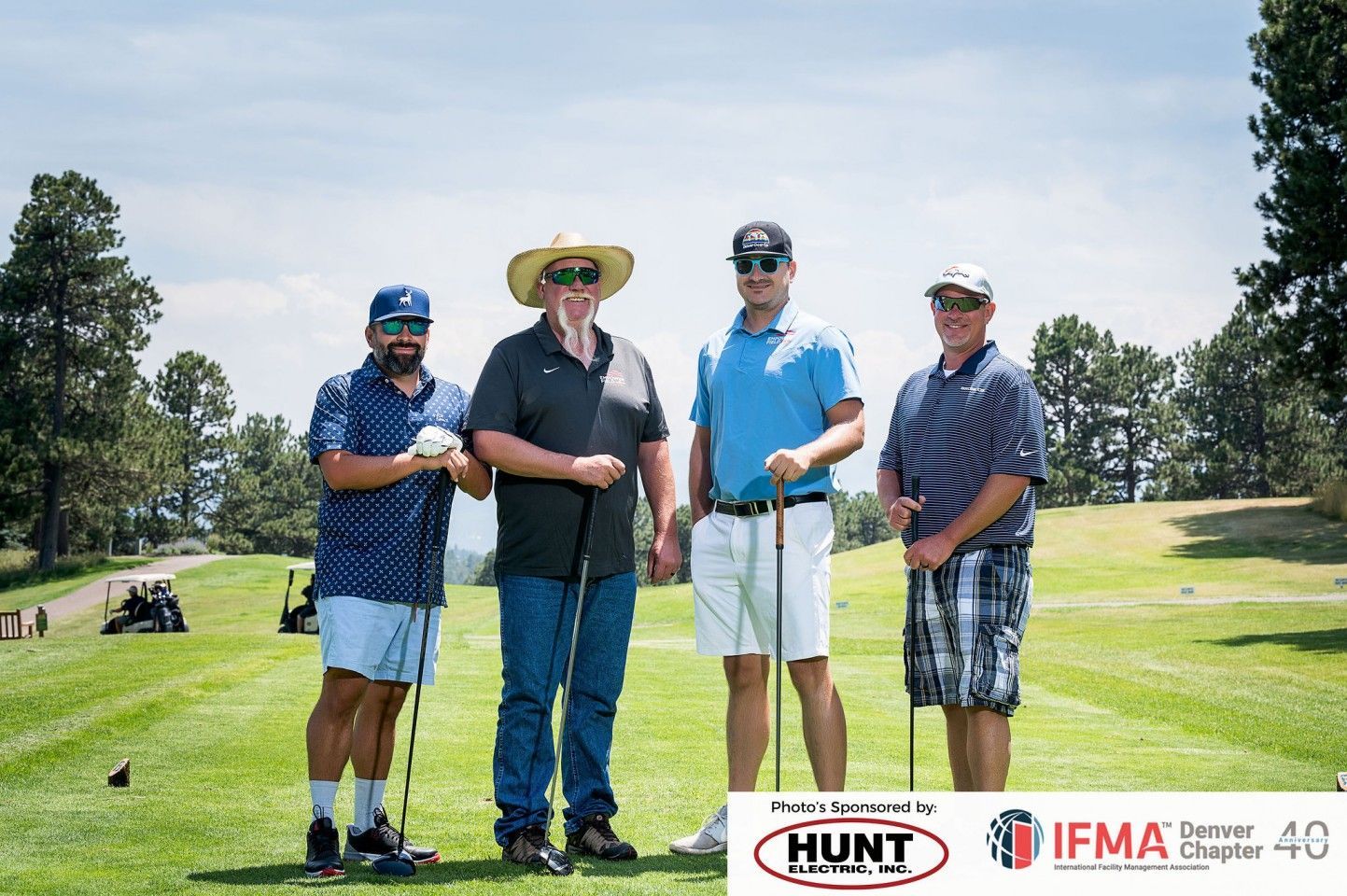 Four men on a golf course holding clubs, smiling. Sunny day, green grass, trees.