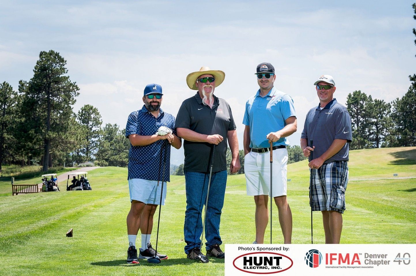 Four men on a golf course posing with clubs. Sunny day, green grass, and trees in the background.