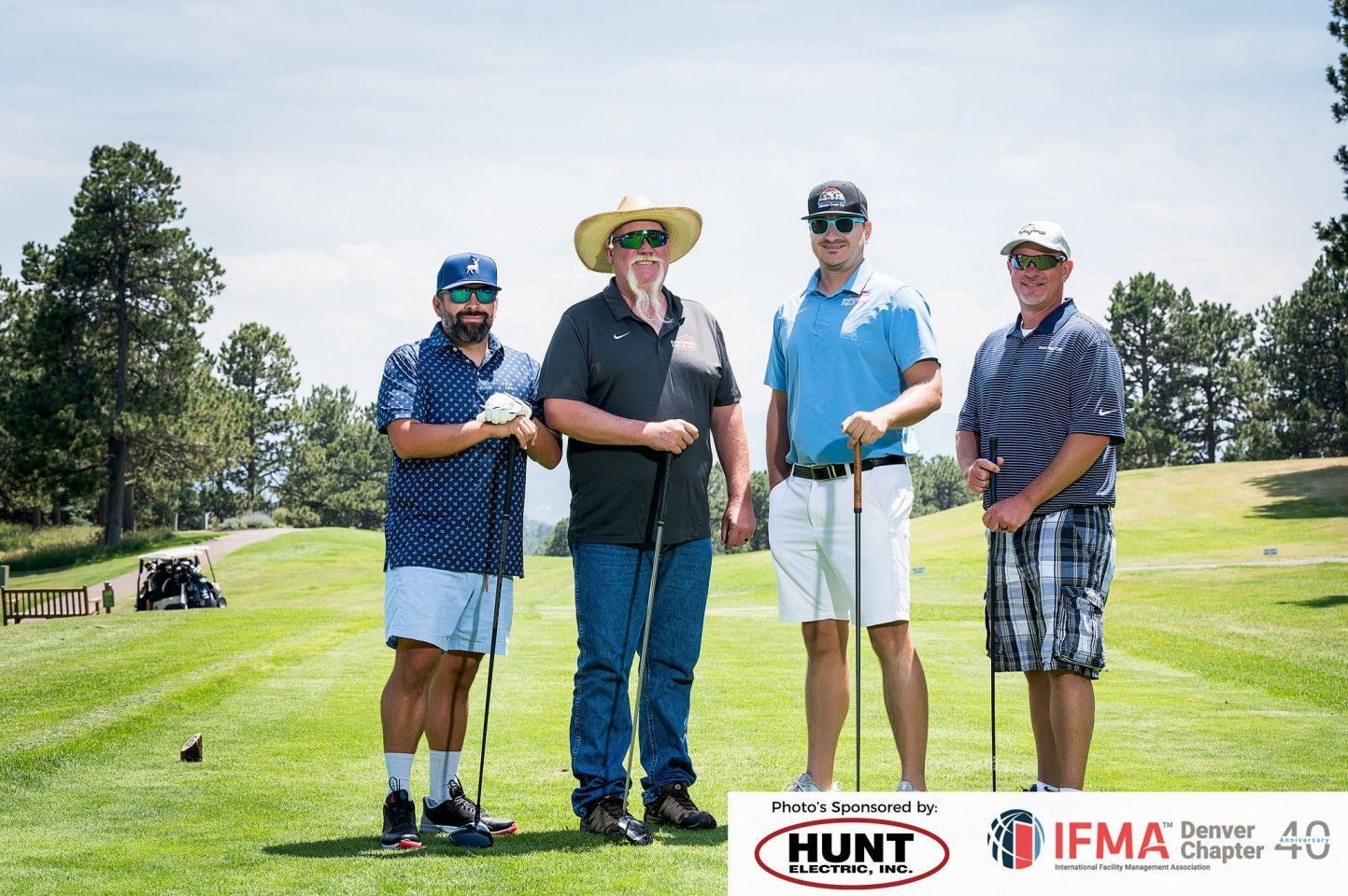 Four men on a golf course pose with clubs. Sunny day, green grass, two men wear hats, all smiling.