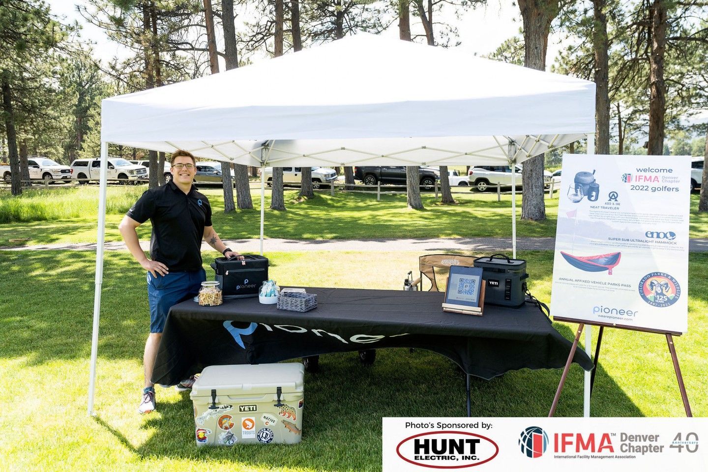 Man stands at a white tent with a table displaying items. A sign is to the right. Outdoors on grass.