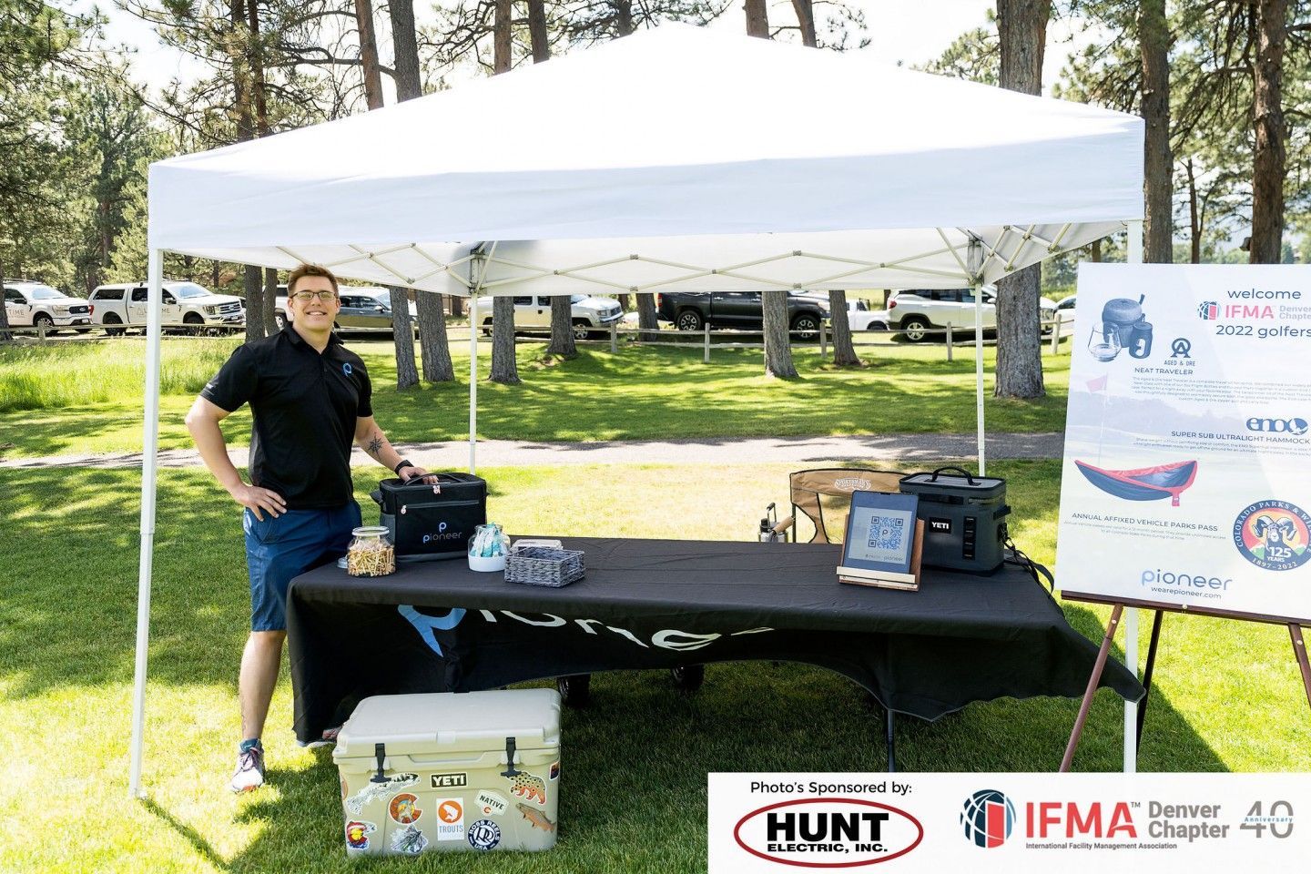 Man stands behind a table under a white tent at an outdoor event. Signage is visible to the right.