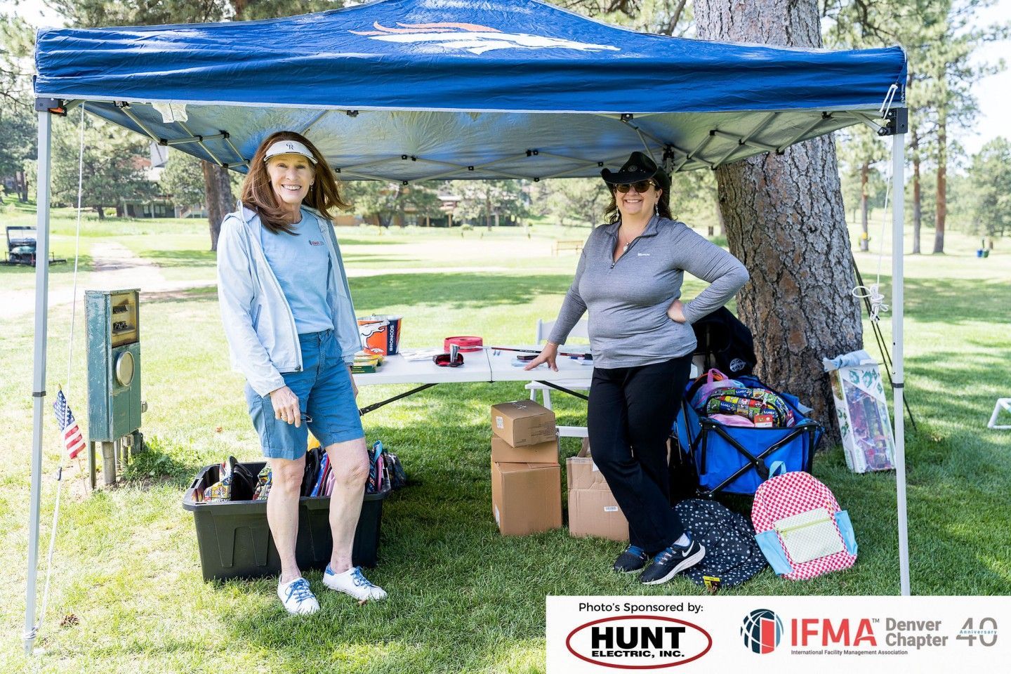 Two women at a table under a blue canopy at an outdoor event. Boxes, bags, and items for sale are on display.