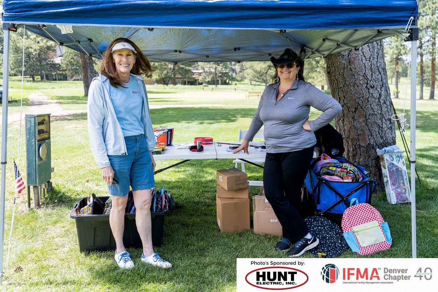 Two women stand at a table under a blue canopy in a park. Items and boxes are on the table.