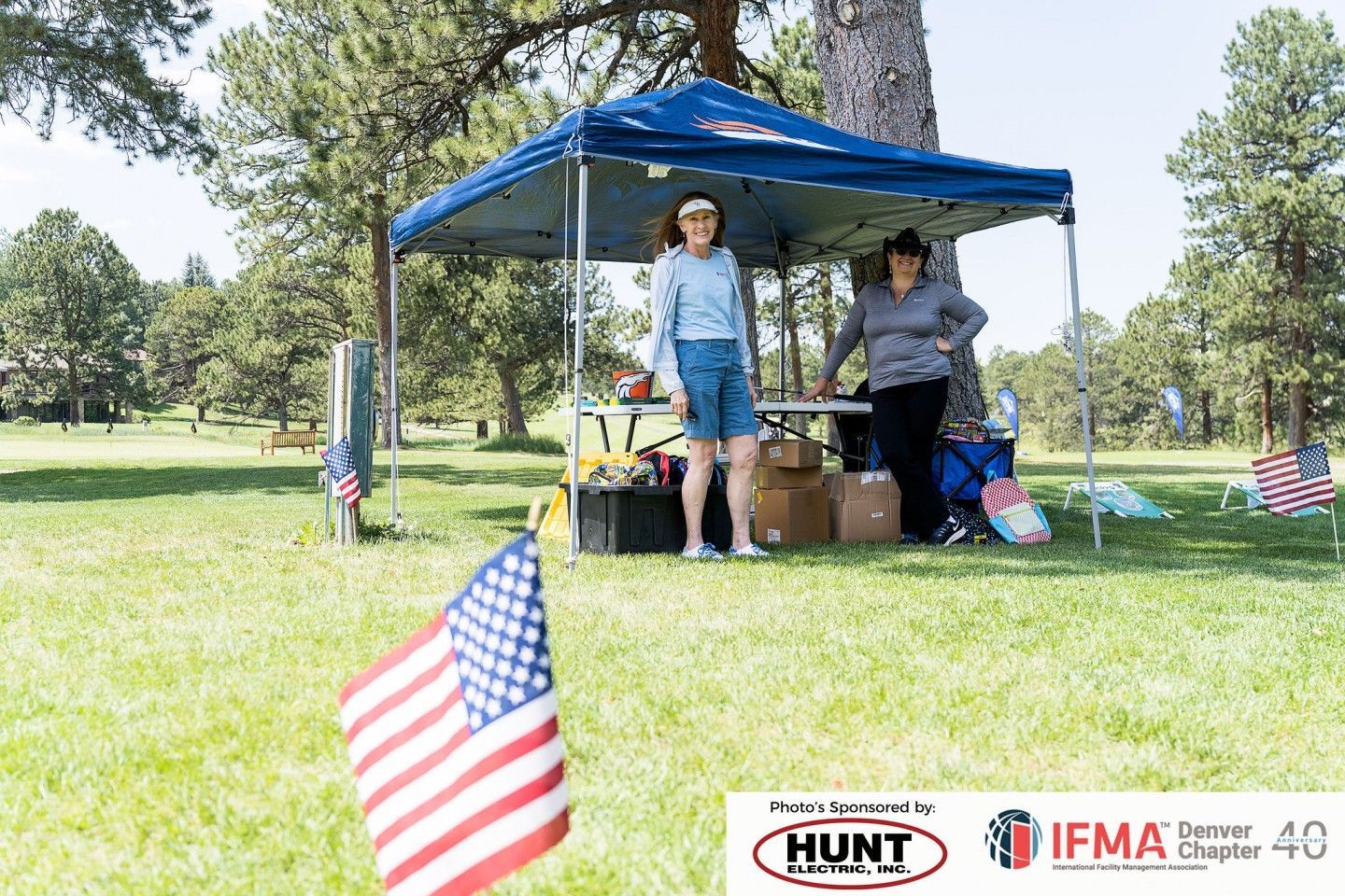 Two people at a table under a blue canopy in a park, with an American flag in the foreground.