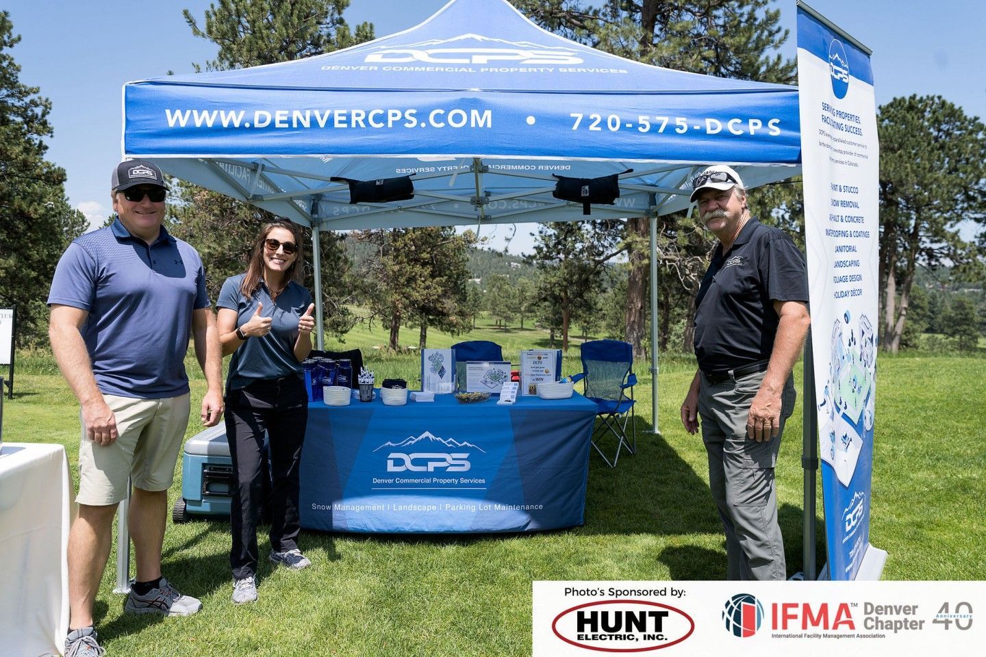 Three people stand at a Denver CPS booth at an outdoor event. A banner above says www.denvercps.com.
