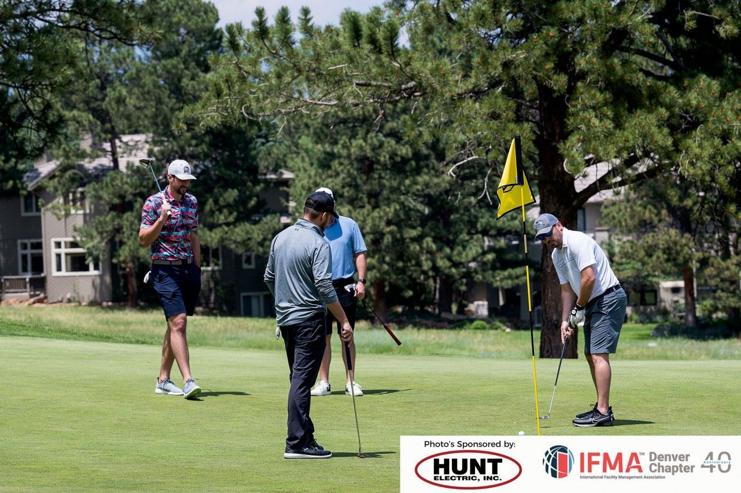 Four golfers on a green; one putts, others watch. Trees, flag, and a building are in the background.