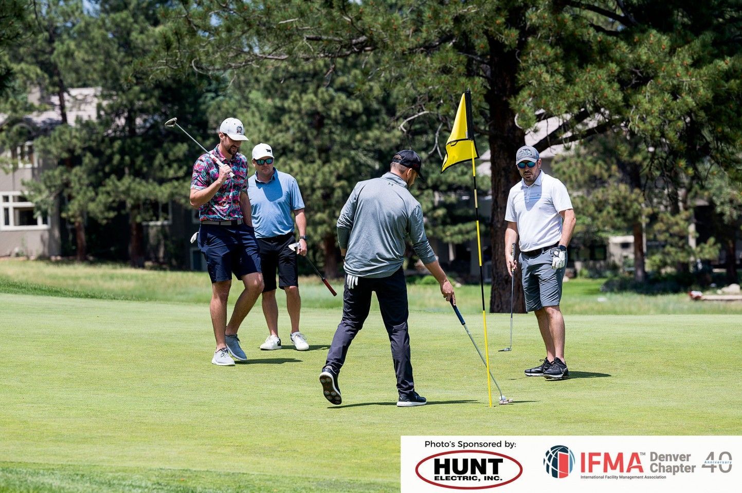 Men golfing: one lines up a putt as others watch, sunny day on the green with a flag.
