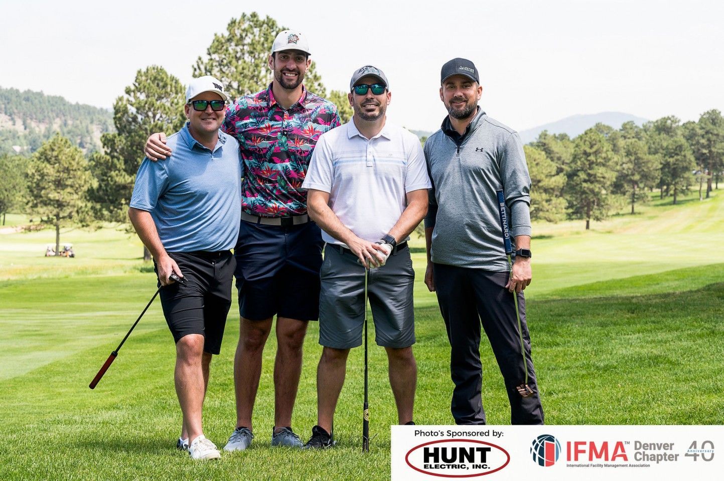 Four men smiling, holding golf clubs on a green course, sponsored by Hunt and IFMA.
