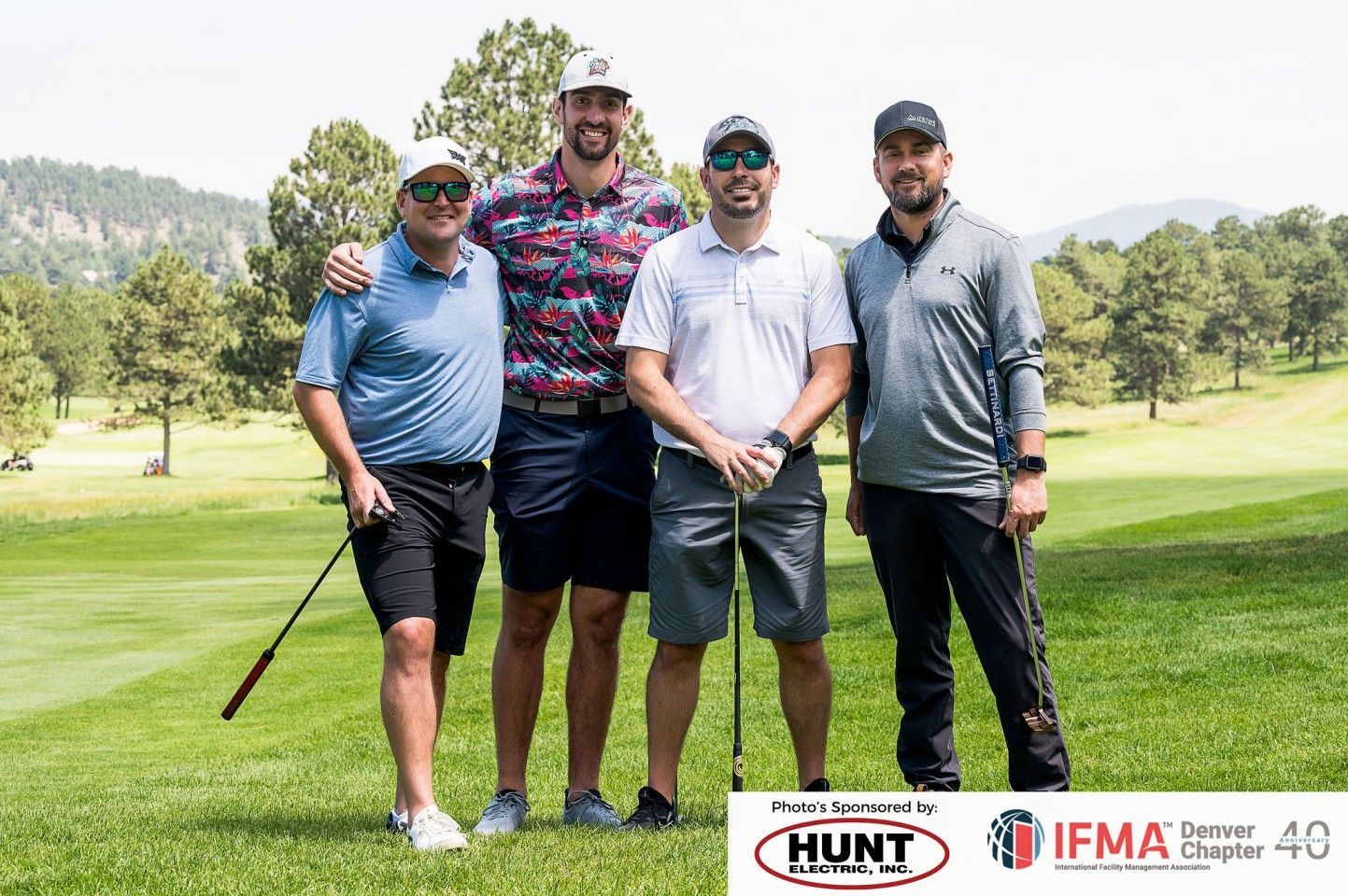 Four men on a golf course pose for a photo, arms around each other. Green grass and trees are in the background.