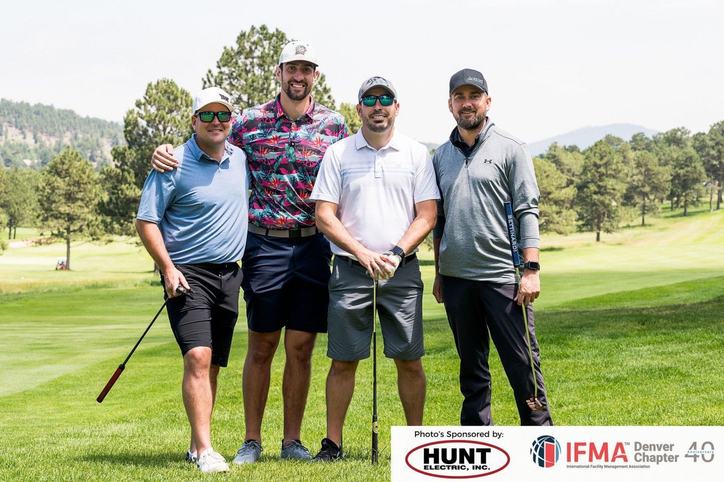 Four men standing on a golf course, smiling, holding golf clubs. Sunny day.