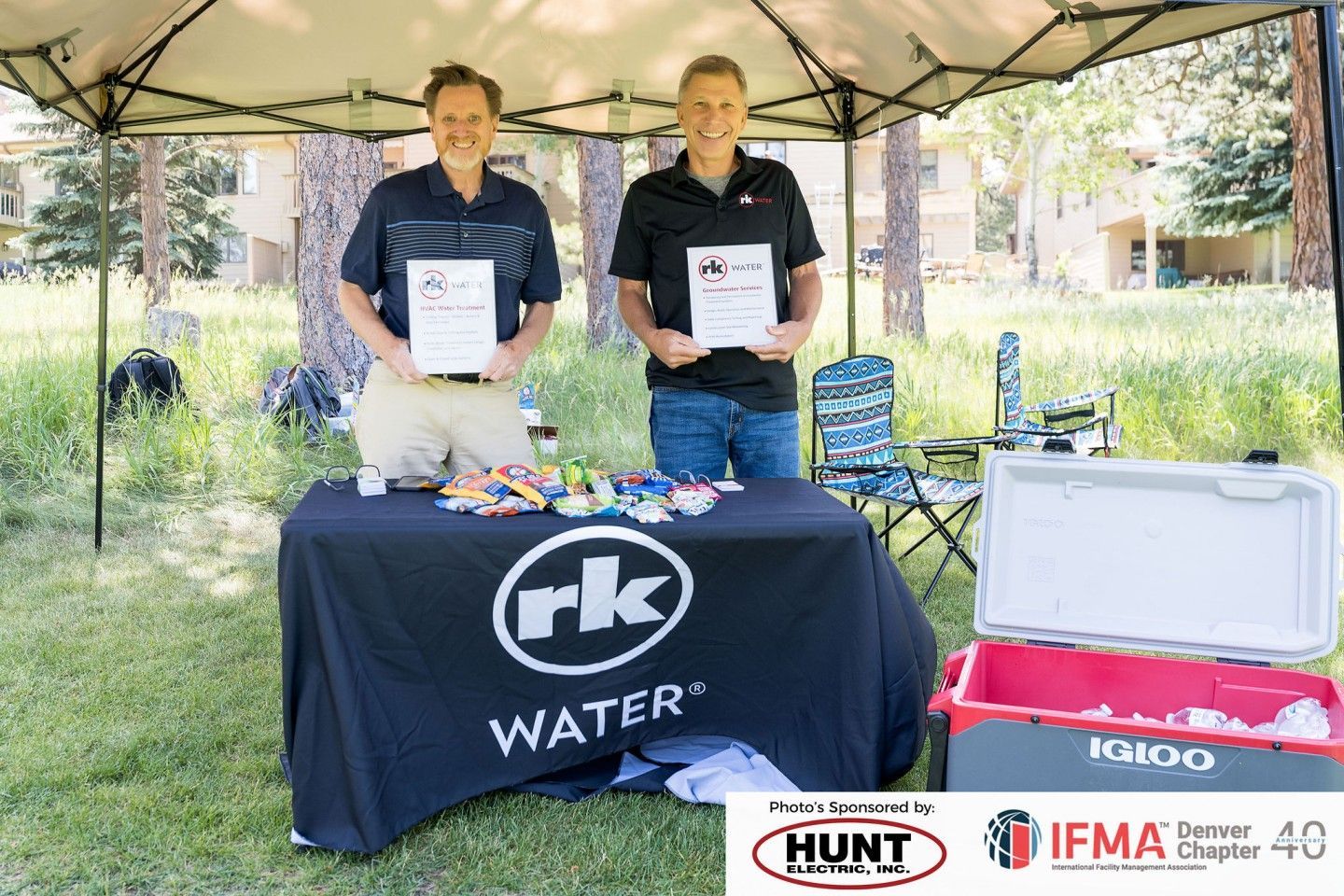 Two people stand behind a table with water-related products under a tent. They hold papers and smile.