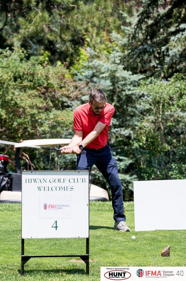 Man in red shirt swings a golf club on a green course, sign reads 