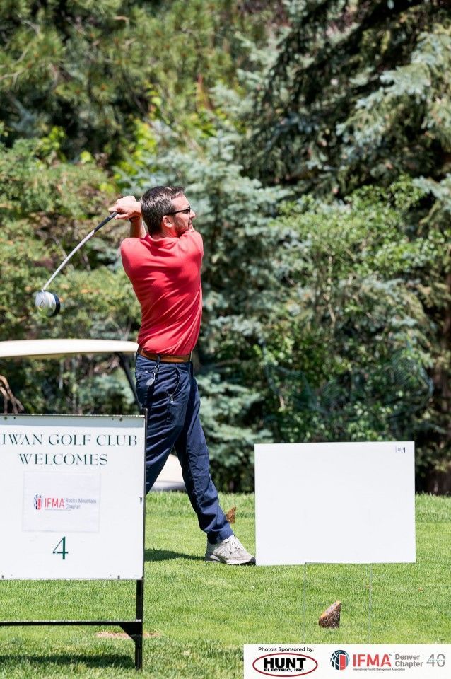 Man in red shirt swings a golf club on a course; trees in the background, a tee marker.