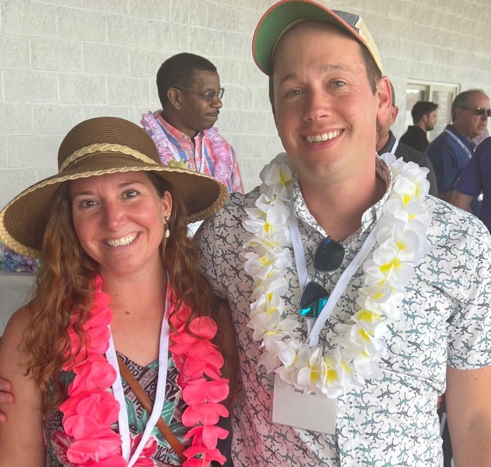 A smiling couple with leis pose outdoors. A man wears a patterned shirt and a hat; the woman wears a wide-brimmed hat.