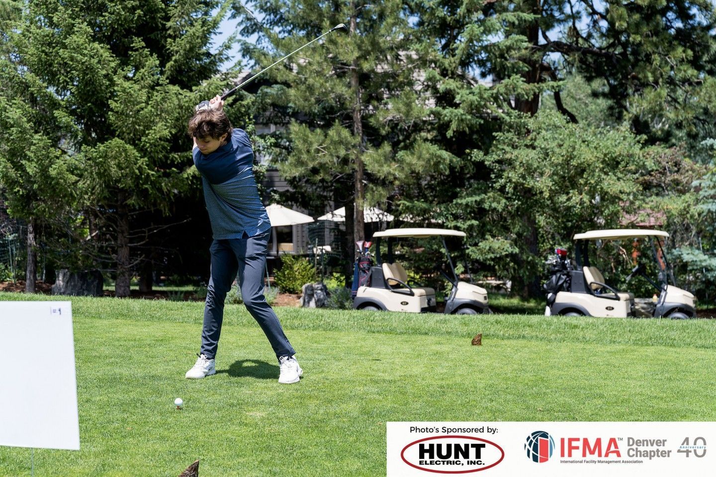 Golfer swings club on a green lawn; golf carts and trees in the background. Sunny day.