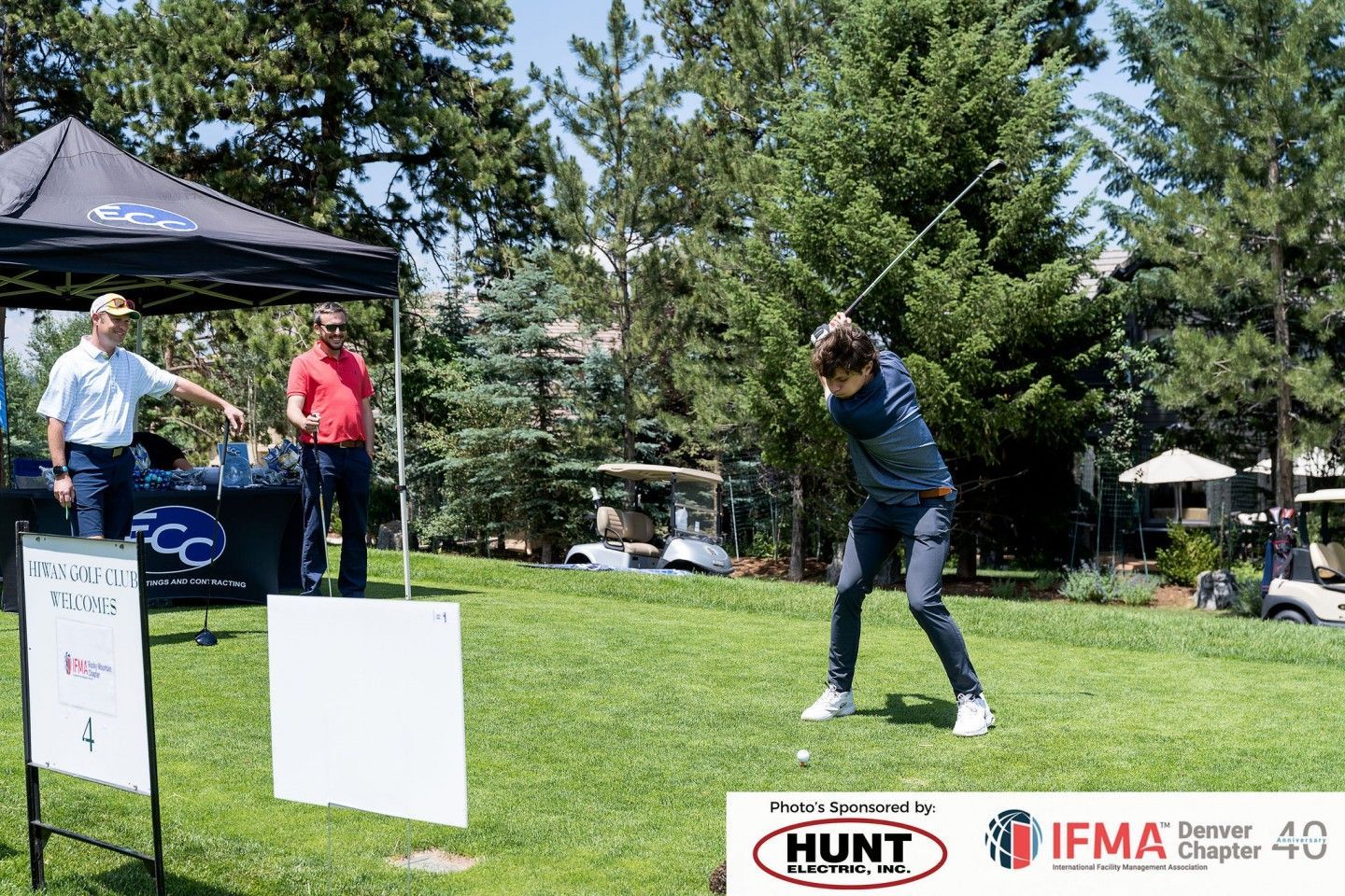 Man swings golf club on a green, two men watch near a tent.