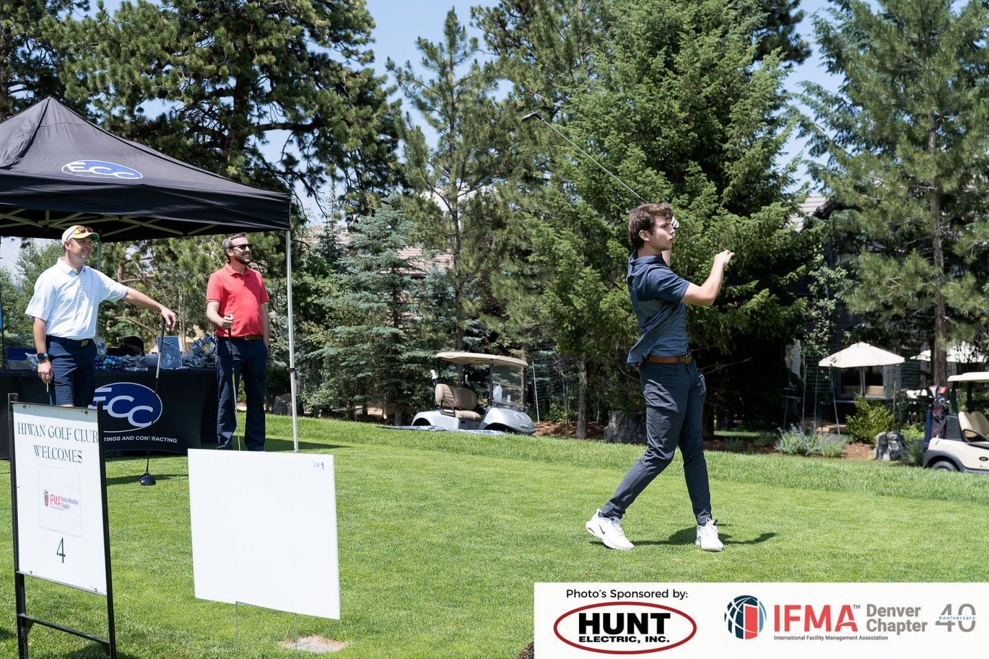 Man swinging a golf club on a green field. Two people watch near a tent; golf carts and trees in background.