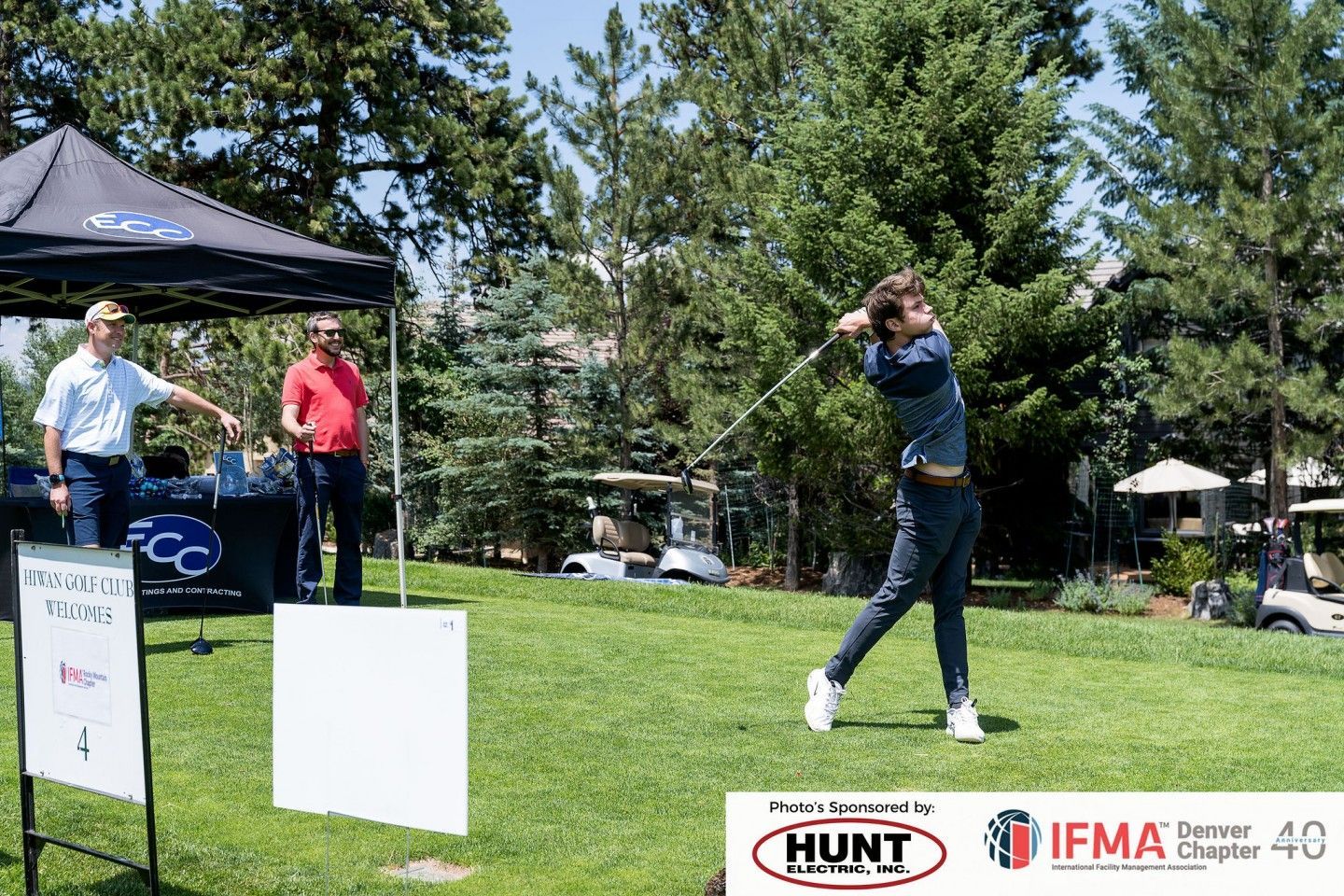 Man swings a golf club on a green course; two men stand nearby.