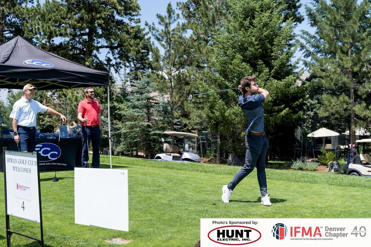 Man swinging a golf club on a green lawn, near a tent and trees on a sunny day.