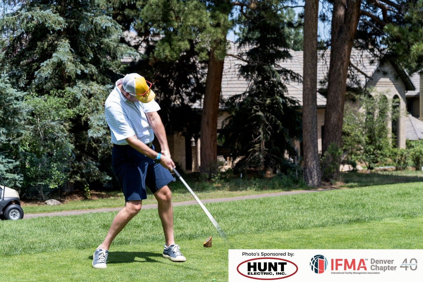 Man swings a golf club on a green lawn, wearing a white shirt and blue shorts, sunny day.