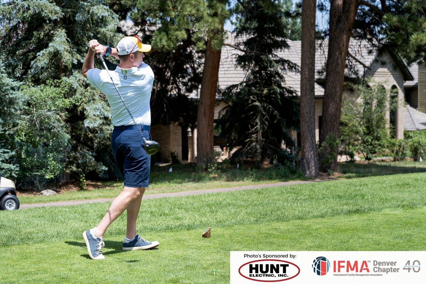Golfer swings club on green; blue shirt, shorts, cap. Sunny day, golf course, house in background.