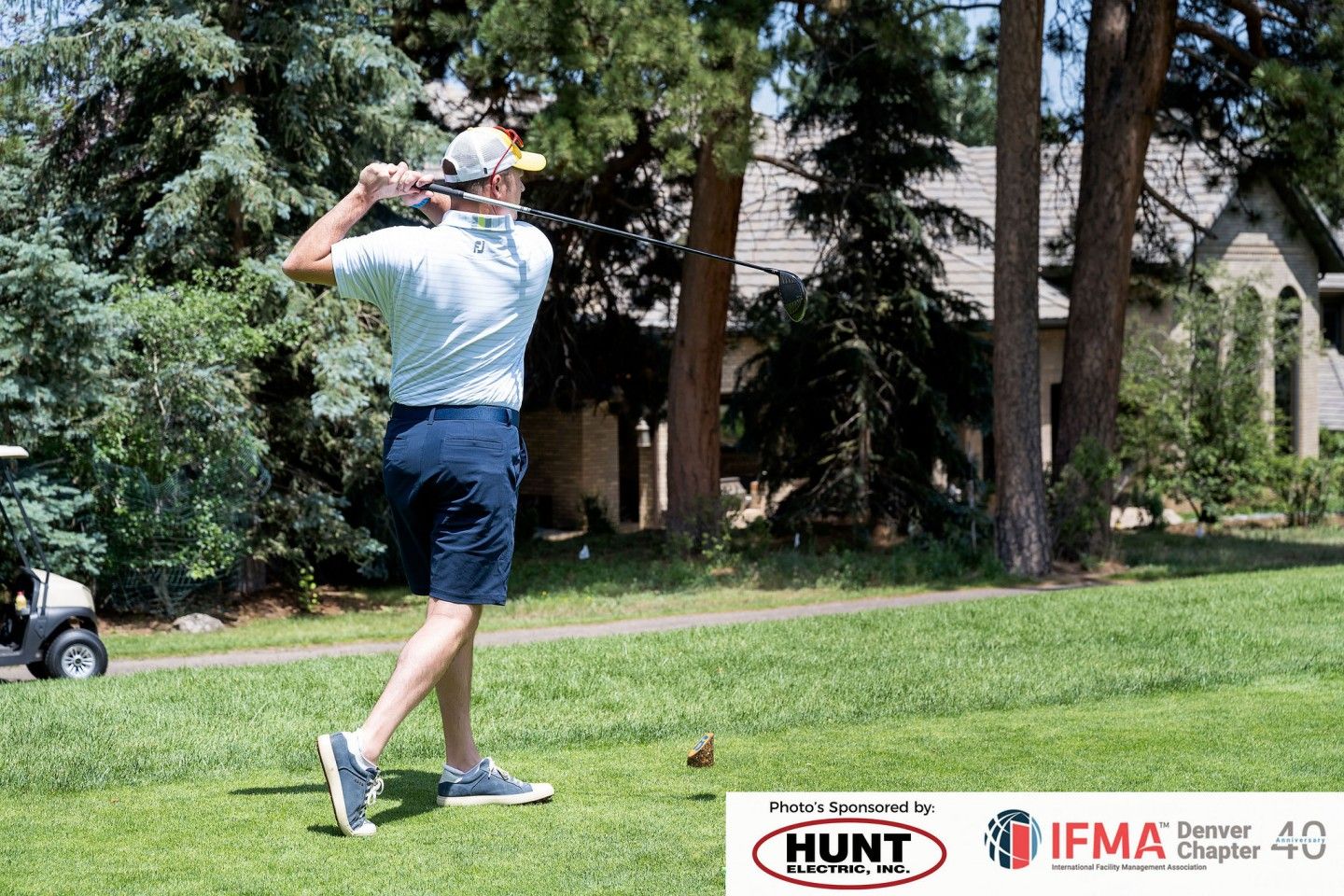 Golfer swinging a club on a green course, trees and a house in the background. Sunny day.