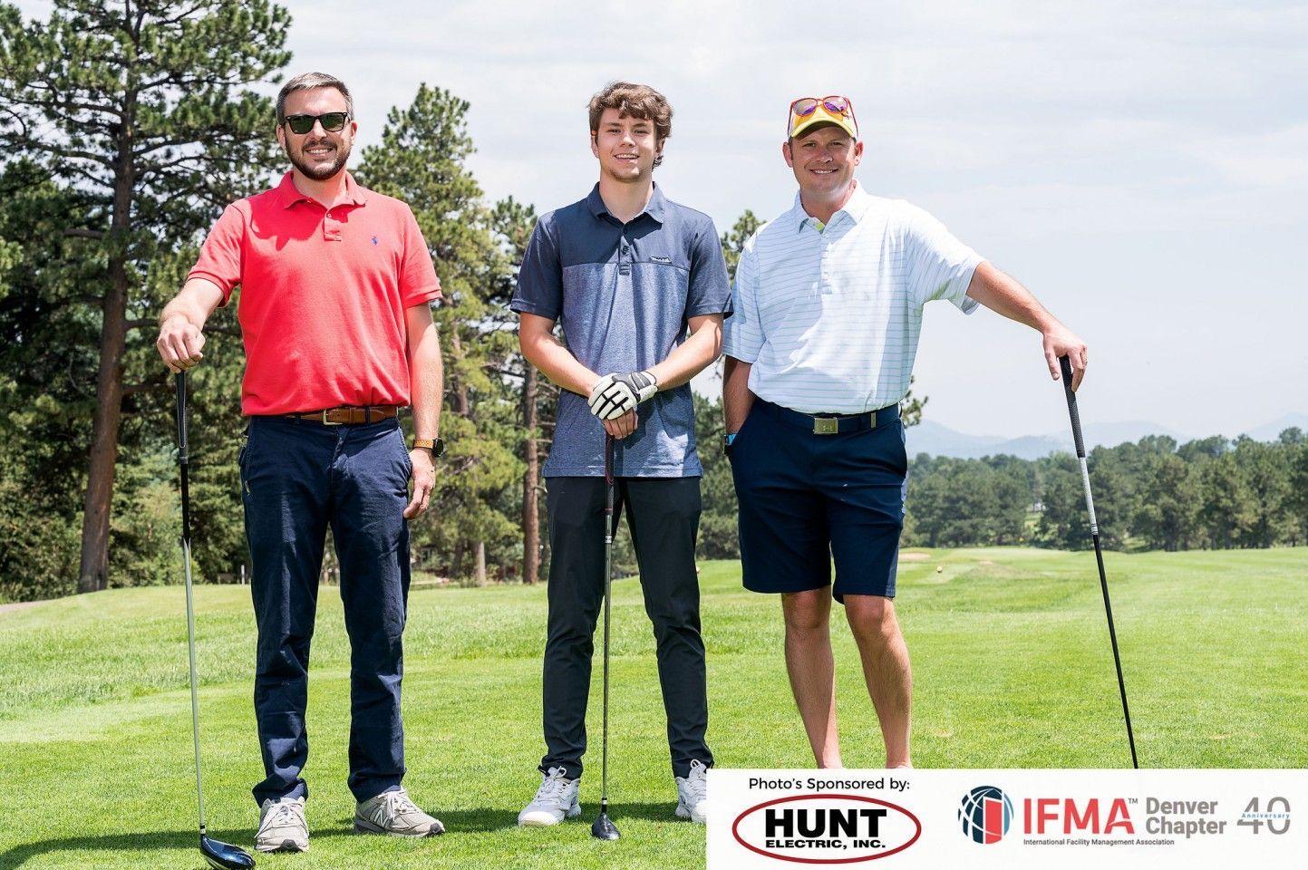 Three men on a golf course pose with clubs. Two in polos, one in red, one in blue. Sunny day.