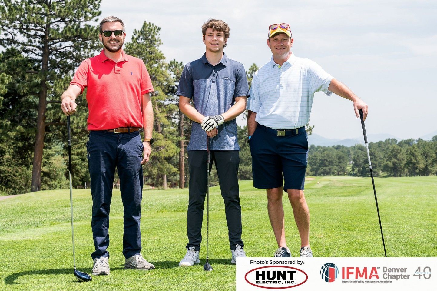 Three men stand on a golf course, holding clubs. Sunny day.