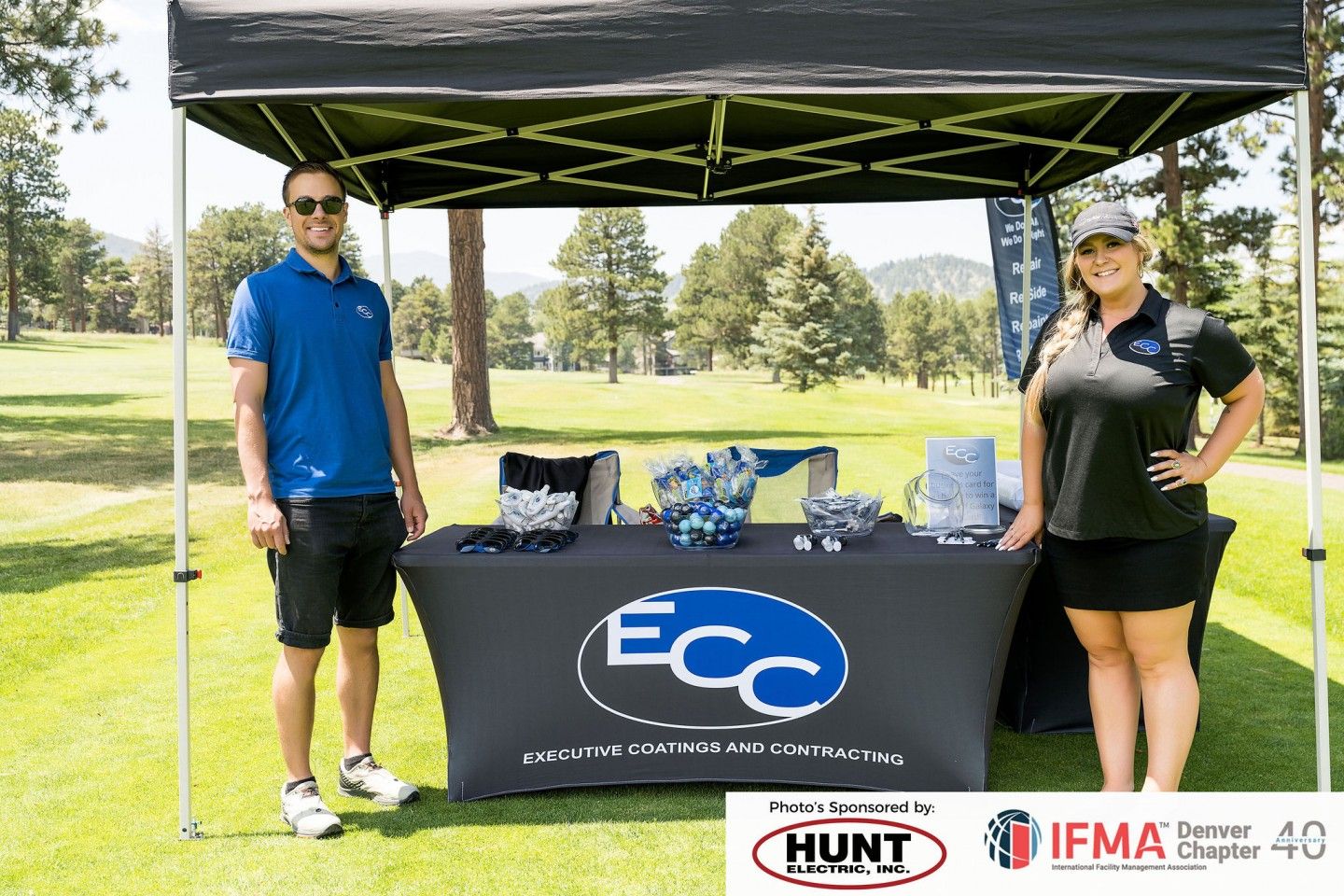 Two people stand by a table under a canopy at a golf event. The table has an 