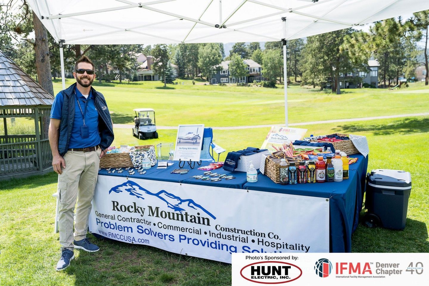 Man standing at a Rocky Mountain Industries booth with logo on table, green golf course setting.