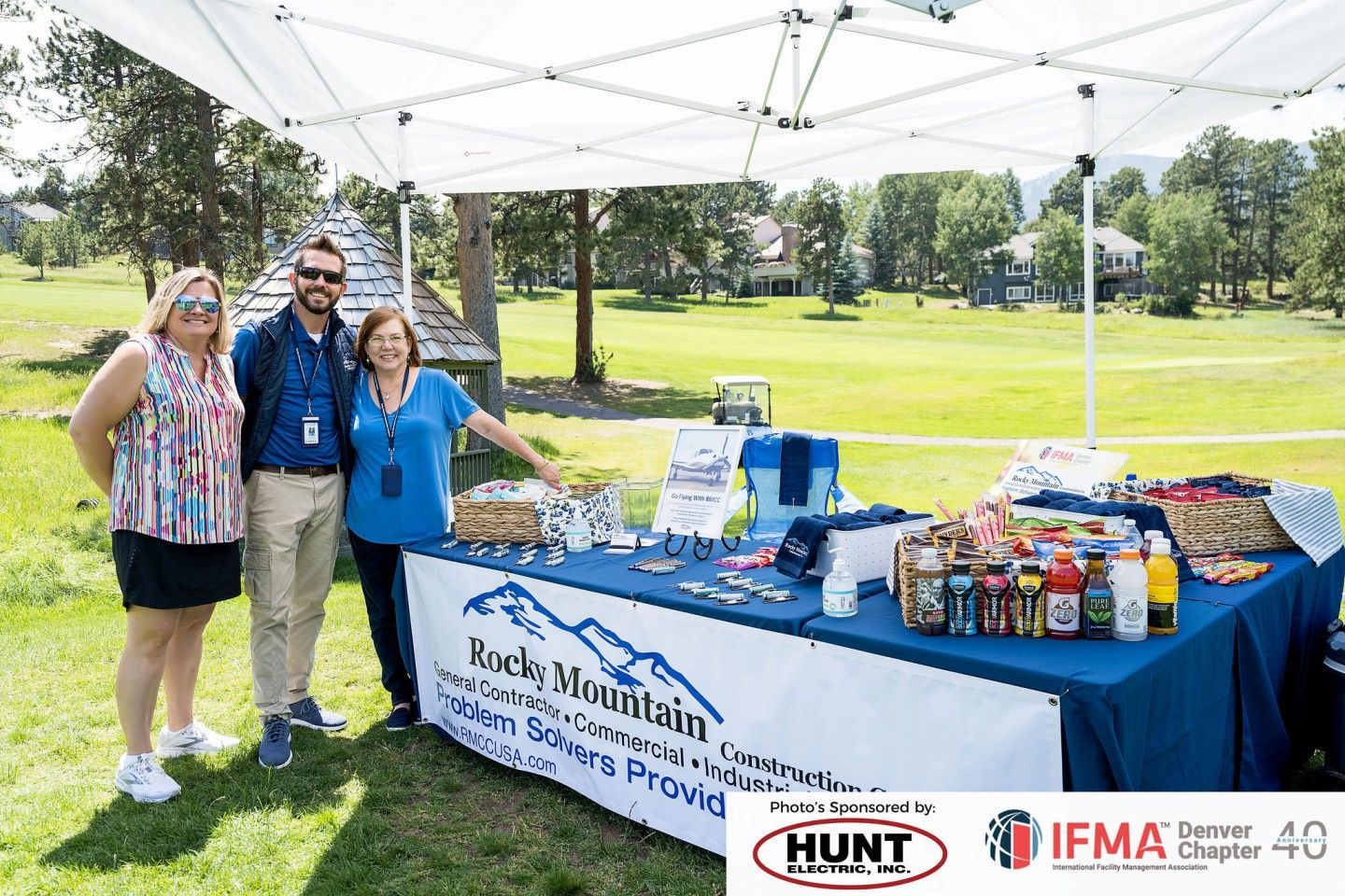 Three people stand behind a table under a canopy at an outdoor event,