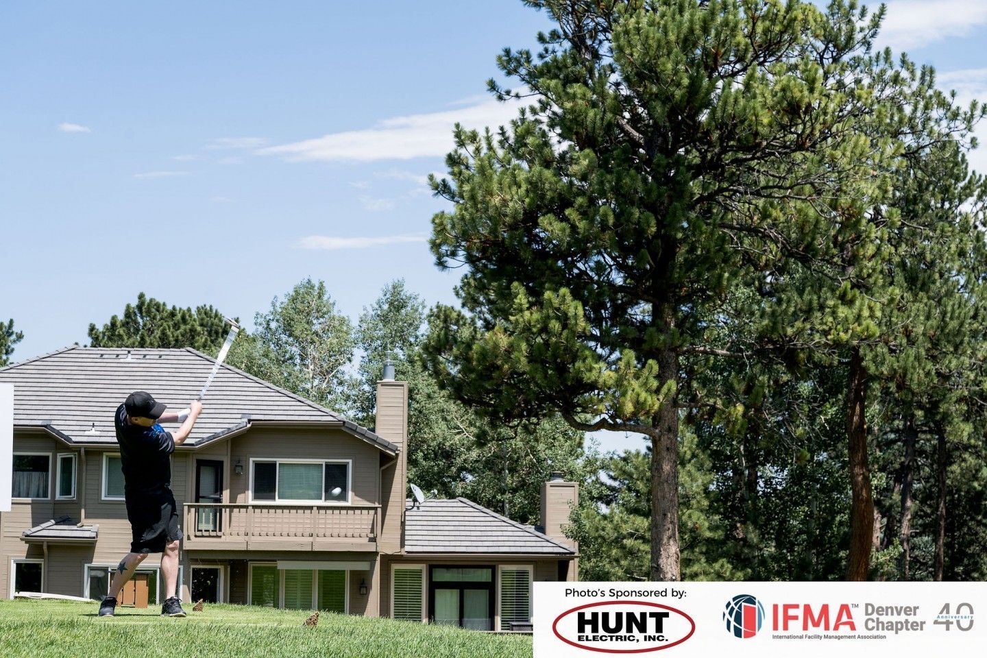 Golfer swings club on a sunny golf course near a large house with trees.